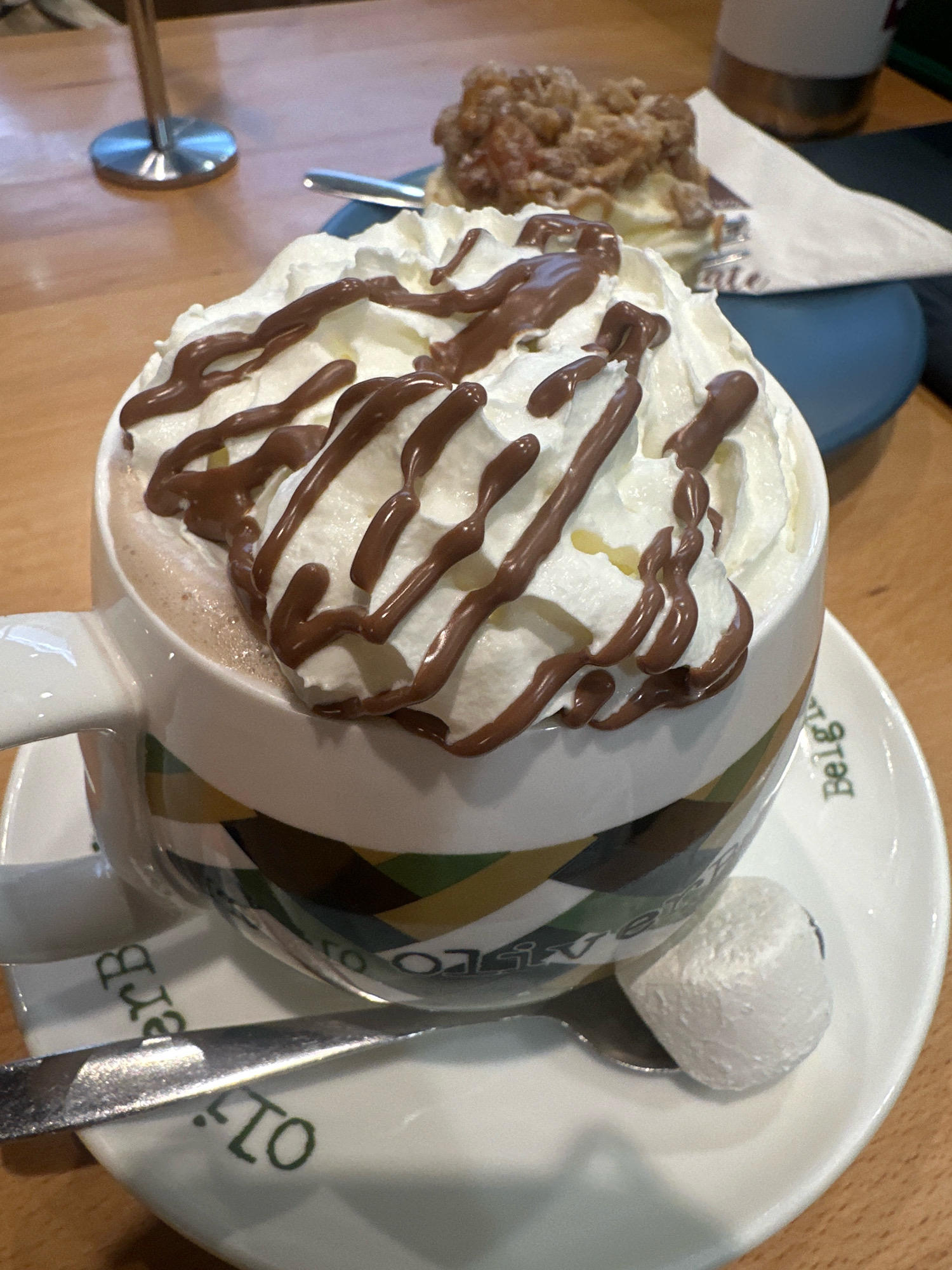 A close-up, eye-level shot captures a mug of hot chocolate topped with whipped cream and chocolate drizzle, sitting on a saucer with a spoon and a marshmallow. The mug, white with a colorful geometric pattern, is the primary subject. The saucer, also white, bears green lettering that appears to be a brand name, possibly related to Belgian food or chocolate. The background reveals a wooden table, a portion of another dessert, and a blurred metal stand, suggesting a cafe or restaurant setting. The overall lighting is warm and natural. No specific location in Coffs Harbour, Australia, can be identified from the visual information provided.