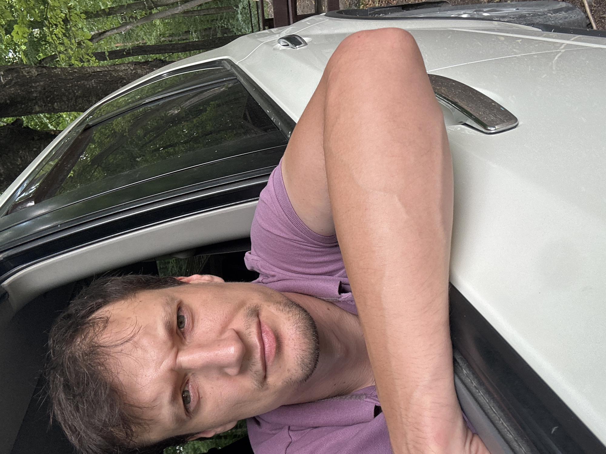 A man with brown hair and a light complexion is partially visible through the open window of a white vehicle, leaning his right arm on the car door. He is wearing a short-sleeved purple polo shirt. His expression is neutral, with his head tilted slightly and his gaze directed towards the camera. The background suggests an outdoor, wooded environment, with trees and foliage visible behind the vehicle. The image appears to be a self-taken photograph, likely a selfie. The location is identified as Shatava, Ukraine.