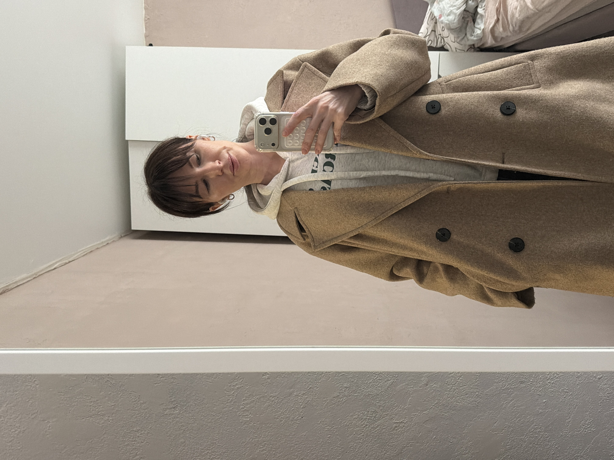 A woman with brown hair is taking a mirror selfie in a room with light-colored walls. She is wearing a light gray hoodie and a long, tan coat with black buttons. A white cabinet is visible behind her. A bed with rumpled bedding is partially visible in the background. The location cannot be determined.
