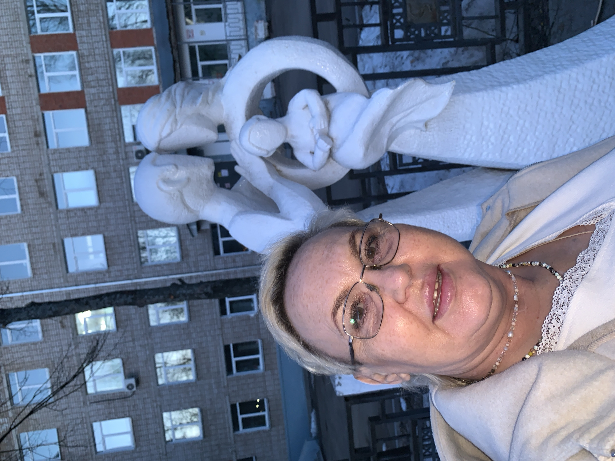 A rotated image captures a smiling woman posing in front of a white sculpture and a residential building, likely in Chaikovsky, Russia.
The primary subject is an adult woman with fair skin, short blonde hair, and metal-rimmed glasses, looking directly at the camera with a gentle smile. She wears a light-colored top with a delicate lace trim visible at the neckline, beneath a cream or light-beige jacket. A multi-colored beaded necklace, featuring various small beads and a larger, lighter, pearl-like bead, adorns her neck.
Behind the woman, dominating the middle ground, is a stylized white sculpture. It depicts a family, with two adult figures seemingly embracing or supporting a smaller child figure nestled within a circular element at their center. The sculpture has a smooth, abstract form, conveying a sense of unity and care.
In the background, a multi-story building with a brick-like facade and numerous rectangular windows is visible. The windows have white frames, and some reflect the sky or surroundings. Bare tree branches, characteristic of colder seasons, are present on the left side of the frame, partially obscuring the building. The lighting suggests it is either an overcast day or late afternoon. No visible text is present in the image. The scene combines personal portraiture with public art and urban architecture, imbued with a pleasant, calm atmosphere from the woman's expression.