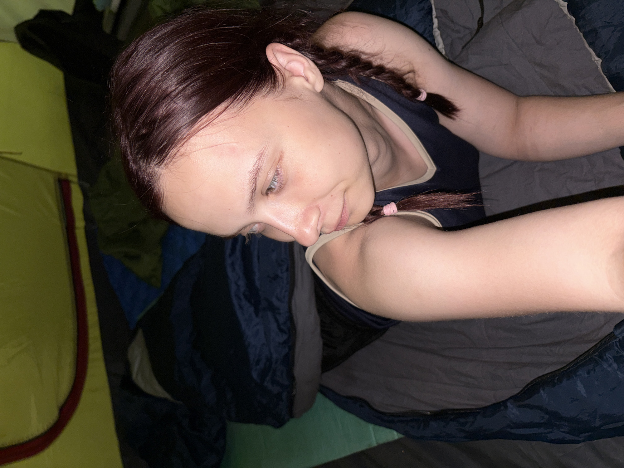 A young woman with dark, braided hair is lying in a sleeping bag inside a yellow and red tent. The sleeping bag is mostly dark gray and dark blue. The inside of the tent is dark. No information to identify the location of the image as Khvalynsk, Russia.