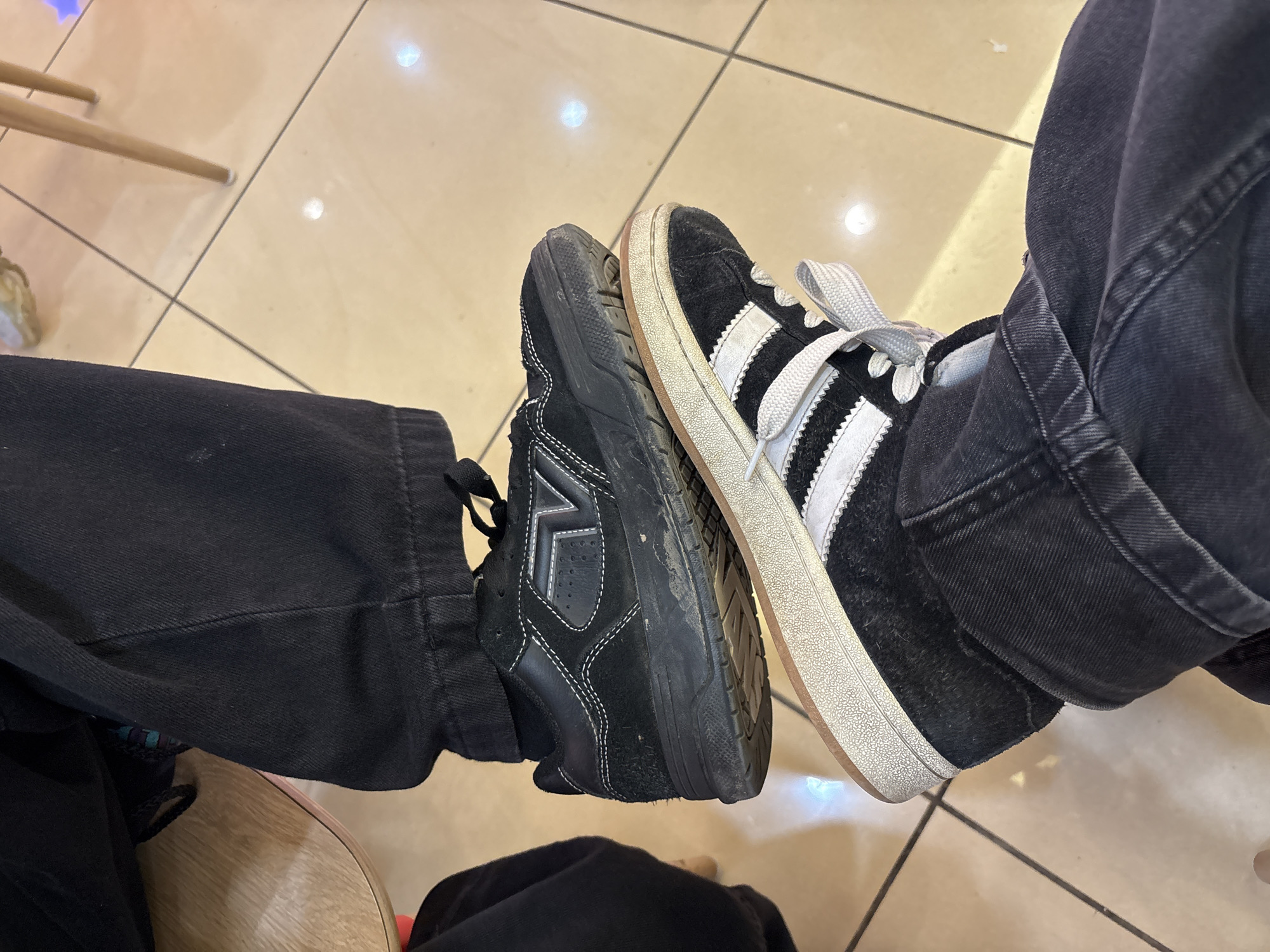 The image shows two pairs of legs wearing black jeans and different black and white shoes. One shoe is an Adidas-style sneaker with white stripes. The other shoe is a black suede sneaker with white stitching and a different logo. The shoes are touching each other. The background appears to be a tiled floor with light reflections. The location is not discernible in the image, and therefore, cannot be confirmed to be in Pleven, Bulgaria.