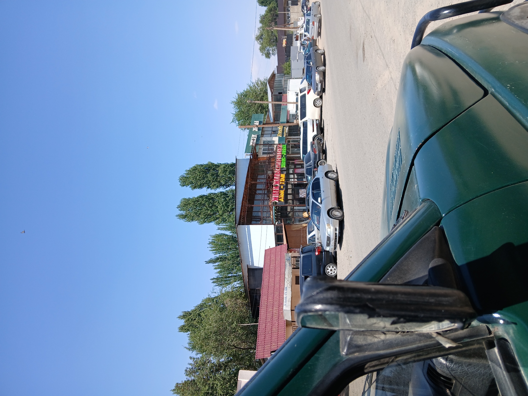 The image captures a street scene in Koktal, Kazakhstan, on a sunny day. In the foreground, the partial view of a dark green vehicle's hood and side mirror is visible, suggesting the photo is taken from inside a car. The street is paved with asphalt and dirt. Several cars are parked along the street and in front of buildings on the right side of the frame. These include a silver sedan, a black minivan, and other vehicles further down.
The background features a row of commercial buildings with various signs, some of which are in a Cyrillic-based alphabet. One prominent building has a red-tiled roof and appears to house a "SUPERMARKET." Another building, with a glass facade, has signs in green and yellow. Tall, slender trees with green foliage line the street behind the buildings. The sky is clear blue, with a few distant birds visible. The overall impression is a typical street view of a town or city in Kazakhstan.