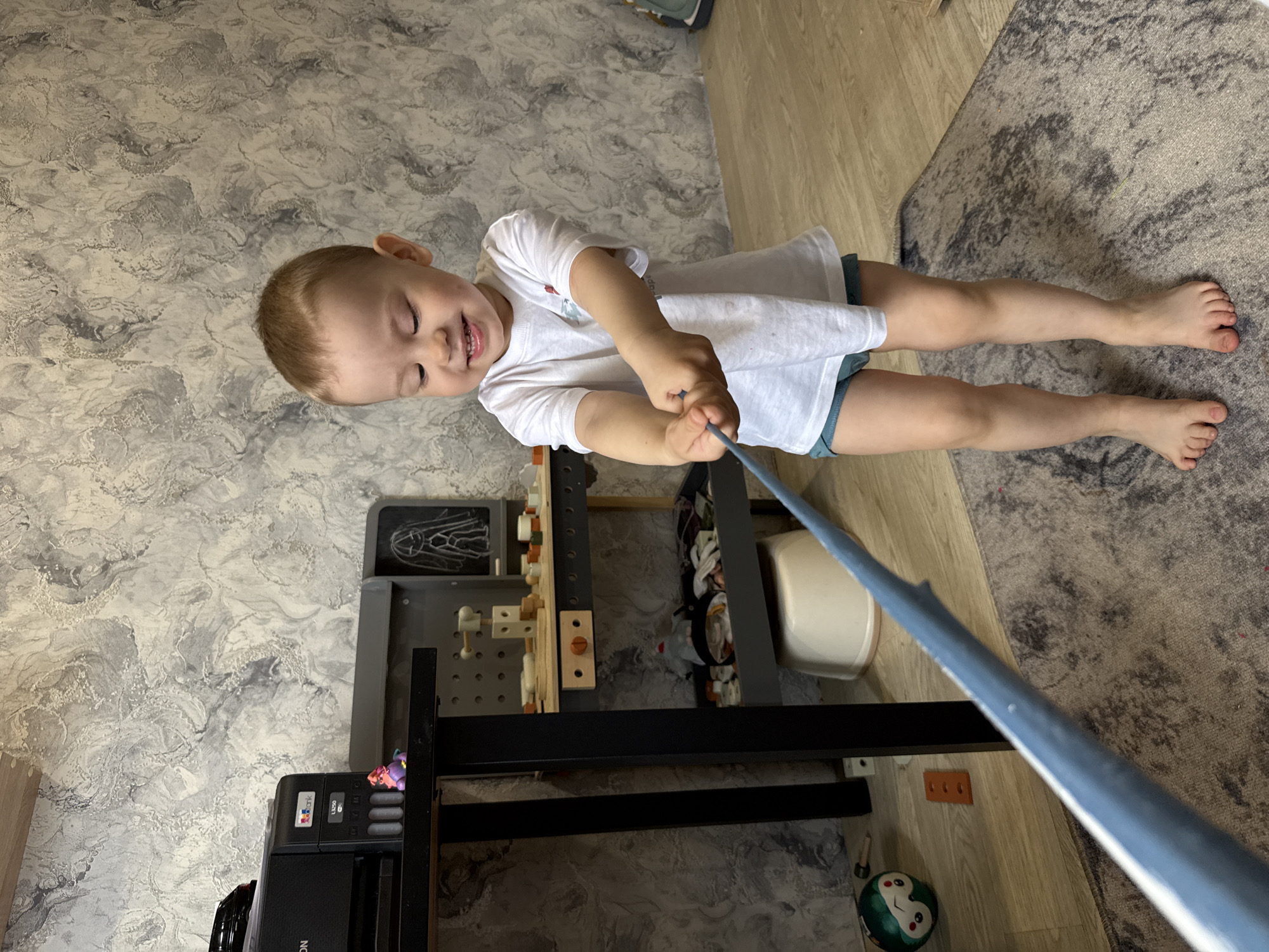A young child with short, light-brown hair, wearing a white t-shirt and blue shorts, is pulling on a blue-colored pole. The child is positioned on the floor, which is covered partly by a gray rug and partly by light-colored hardwood flooring. The floor also features wallpaper with a marbled pattern. In the background, there's a black shelving unit and a white bucket or container. The exact location in the city of Aristovo, Russia, is not discernable from the image.