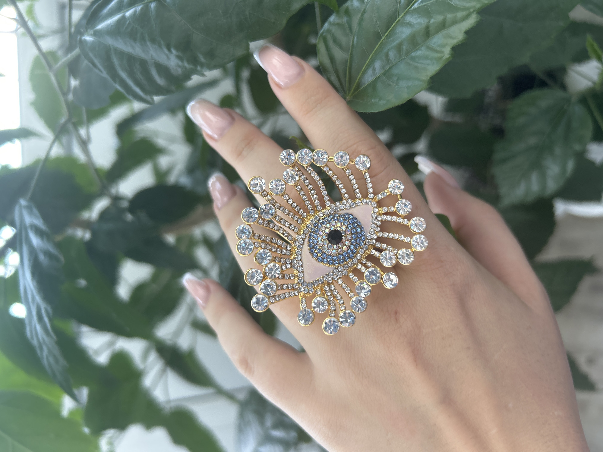 A hand with French manicured nails is adorned with a large, ornate ring on the middle finger. The ring is gold-toned and designed to resemble an eye, with a pink triangular stone at its center, surrounded by smaller blue and black stones that form the pupil and iris. The outer edge of the eye design is embellished with numerous clear, round gemstones set in a radiating pattern, resembling sun rays or eyelashes. The hand is positioned against a blurred background of lush green foliage, suggesting an outdoor or indoor plant setting. The image is a close-up shot, focusing on the ring and the hand. There is no indication of the city of Odesa, Ukraine, within the image itself.