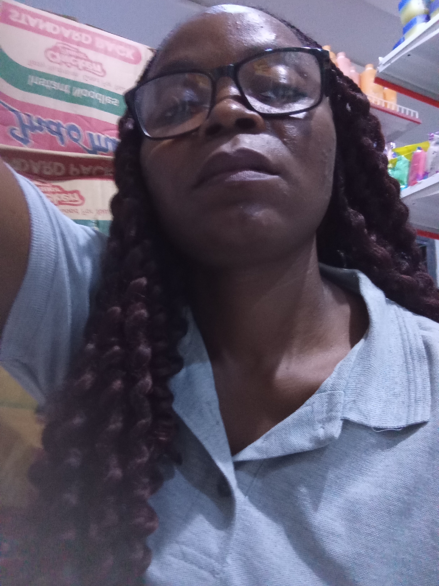 A close-up, high-angle selfie captures a dark-skinned woman with glasses and braided hair. She is wearing a light blue polo shirt. The background is blurry and suggests a retail environment, with shelves of products visible on the right. The blurred signage in the background, though partially illegible, indicates this scene is likely in Howayi, Nigeria. The overall lighting is dim, with some overhead illumination.