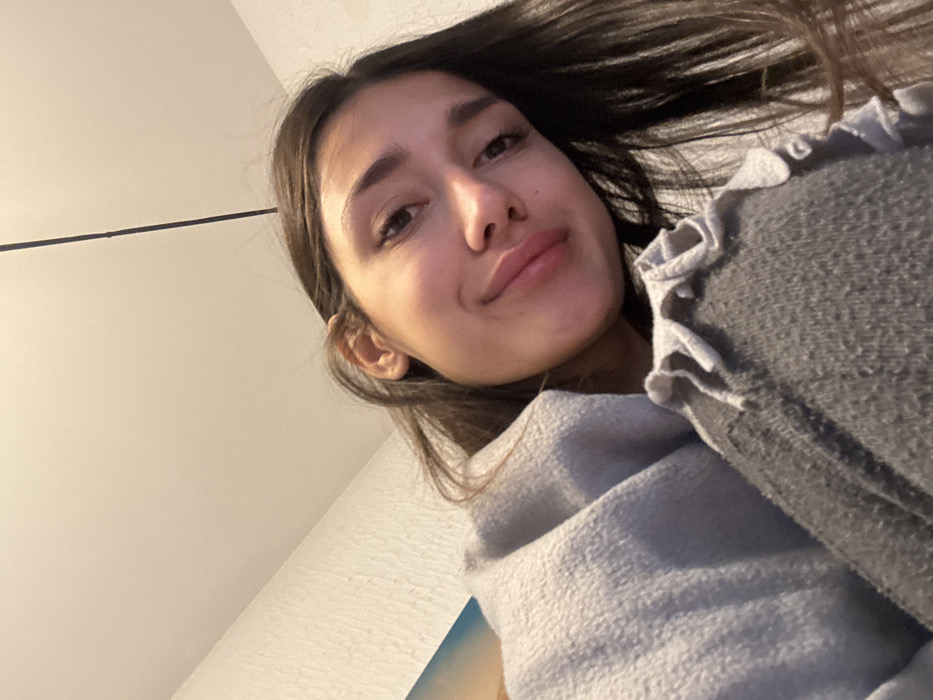 A single female individual is depicted indoors, looking directly towards the camera with a slight head tilt and a subtle upward curvature of the lips. She has long, dark brown hair. Her upper body is covered by a light grey, textured fleece-like garment. A section of dark grey, textured fabric with ruffled edges is visible in the lower right foreground, consistent with clothing or a blanket. The background consists of a light-colored, flat ceiling occupying the upper portion of the frame. A thin, black linear element descends vertically from the ceiling. To the left, a textured, light-colored wall is visible, along with a partial view of what appears to be a colorful picture or abstract art piece featuring blue and orange/yellow hues. This scene is located within a room in Zankovići, Montenegro.
