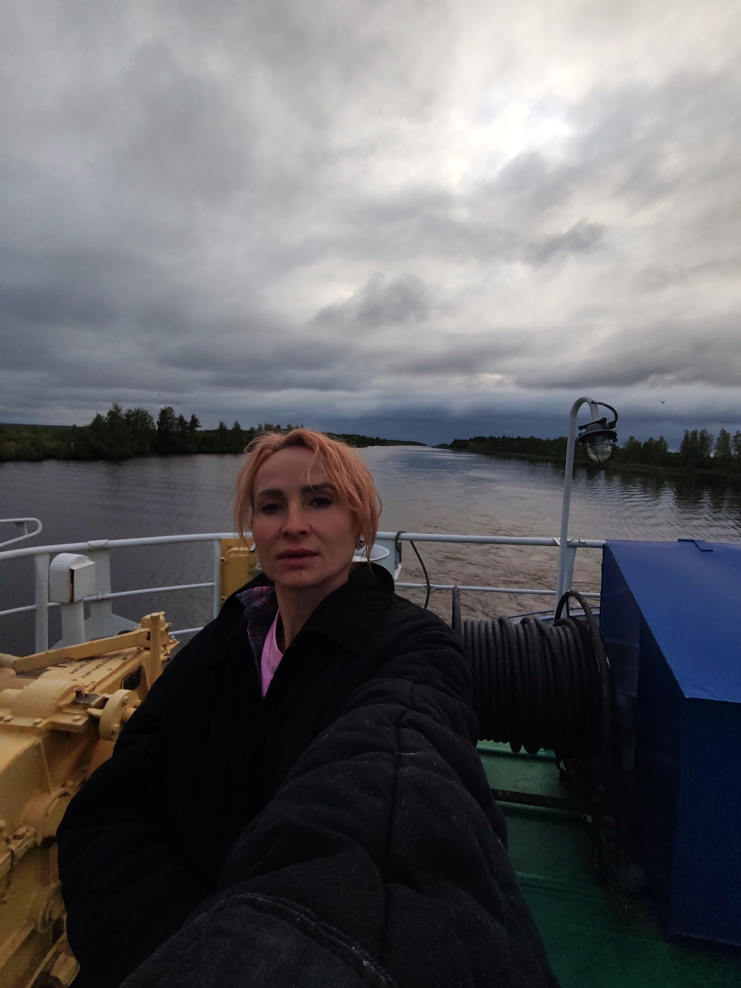 A woman with short, light-colored hair is taking a selfie on the deck of a boat. She is wearing a dark jacket over a pink shirt. The boat appears to be on a body of water, possibly a river or canal, with wooded banks visible in the distance. The sky is cloudy and overcast, suggesting a cool or potentially rainy day. The location is identified as Ozernoe Ust'e, Russia. Other objects visible on the deck include a yellow piece of machinery, a large spool of rope, and a blue structure. The overall atmosphere is somber due to the cloudy sky.