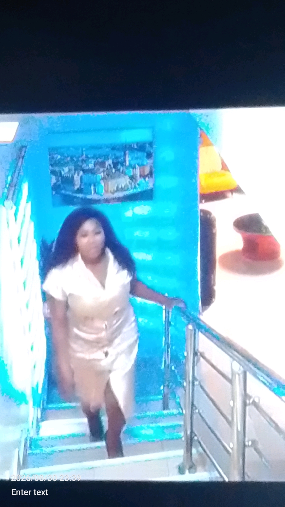A medium-shot image captures a woman standing on a brightly lit staircase with blue walls and a polished metal railing. She is wearing a short, light-colored satin dress with short sleeves and a V-neck. Her dark, wavy hair falls past her shoulders. The woman is looking slightly off-camera to her left, with her right arm resting on the railing. The staircase appears to be indoors, with a painting of a city landscape hanging on the wall to her left. The lighting is strong, casting shadows and creating a vibrant, almost surreal atmosphere. There are no discernible clues within the image to suggest the specific location in Lokoja, Nigeria.