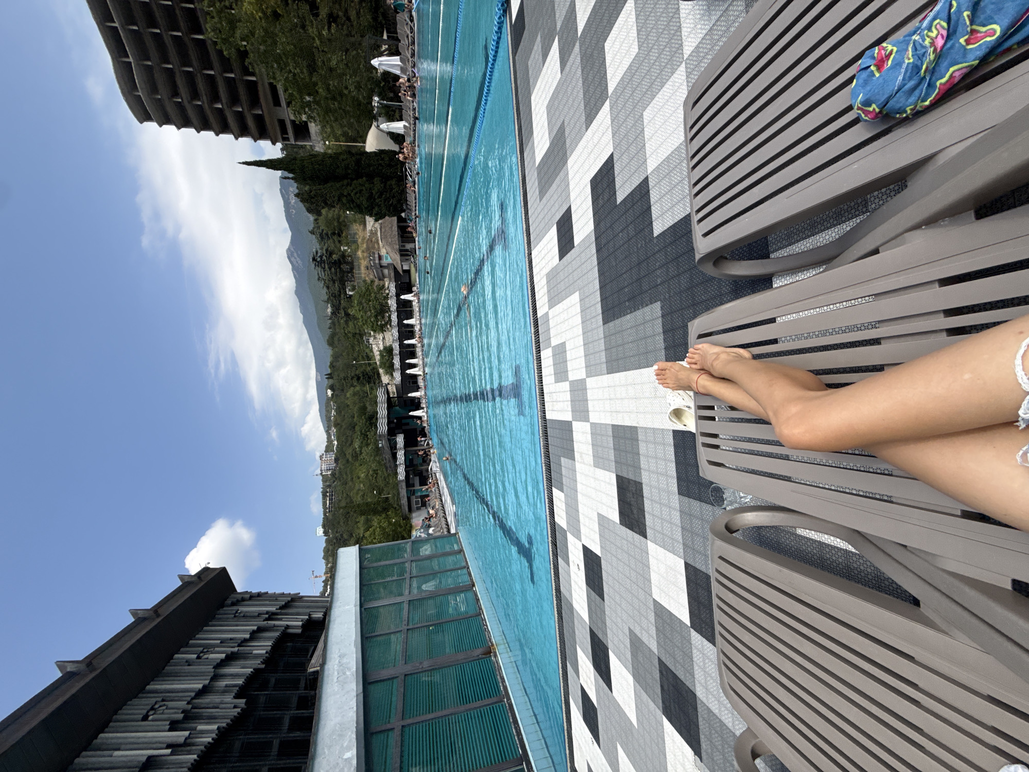 An outdoor Olympic-sized swimming pool is depicted from a first-person perspective, with the viewer's legs resting on a lounge chair in the foreground. The pool is filled with bright blue water and is divided into lanes by white ropes. Several people are visible swimming or relaxing in and around the pool. In the background, a multi-story building with a modern, angular facade is on the left, and a tall, unfinished concrete building is on the right, situated on a lush, green hillside. Mountains form the backdrop, and the sky is a clear blue with a few white clouds. The poolside area is paved with a mosaic of black, white, and gray tiles.