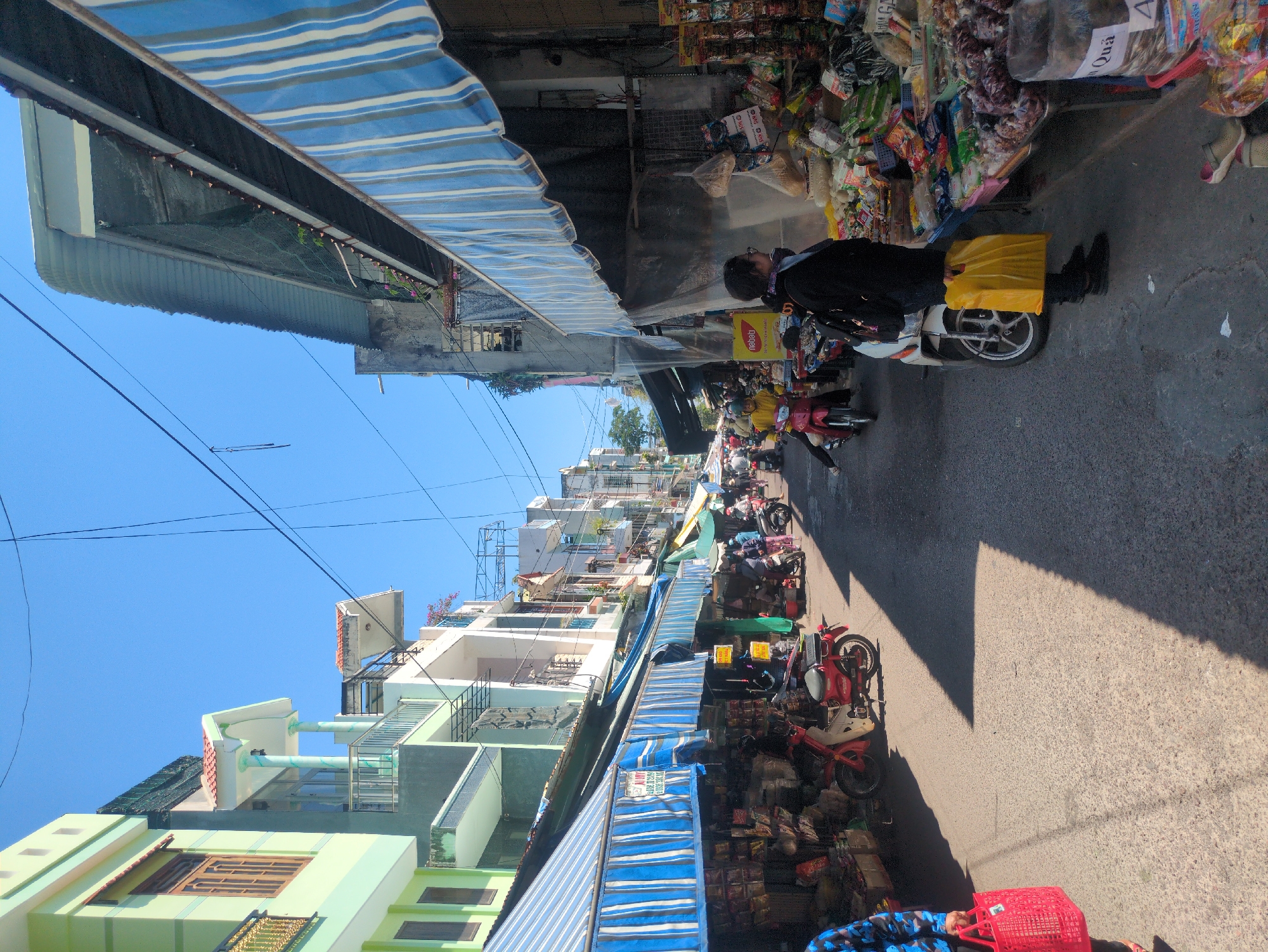 This image depicts a bustling market street in South Quy Nhon Ward, Vietnam, under a clear blue sky. The narrow asphalt street is flanked by multi-story residential and commercial buildings.
On the left side, buildings with light green and white facades feature balconies and ornate railings. Blue and white striped awnings extend from these buildings, covering numerous market stalls. These stalls display a variety of goods, including packaged items and possibly produce. A red Honda Super Cub style motorcycle is parked at the edge of the street near one of these stalls. A person wearing a blue and black patterned top is partially visible in the foreground, carrying a red mesh shopping bag.
On the right side, older, concrete-colored buildings also have blue and white striped awnings, under which more market stalls are set up. These stalls are heavily laden with diverse merchandise, including fruits, vegetables, and packaged goods. A yellow "Nebati" advertisement sign is visible. In the mid-ground, a person wearing a black jacket, dark pants, and glasses stands beside a white scooter, holding a yellow plastic bag, appearing to browse the items on a stall table.
The street itself contains several other motorcycles and scooters, both parked and in motion, indicating active traffic. One individual on a pink scooter, wearing a yellow top, is visible further down the street. Strong shadows are cast across the street from the buildings and awnings, suggesting bright sunlight. Numerous utility wires crisscross overhead against the clear blue sky.