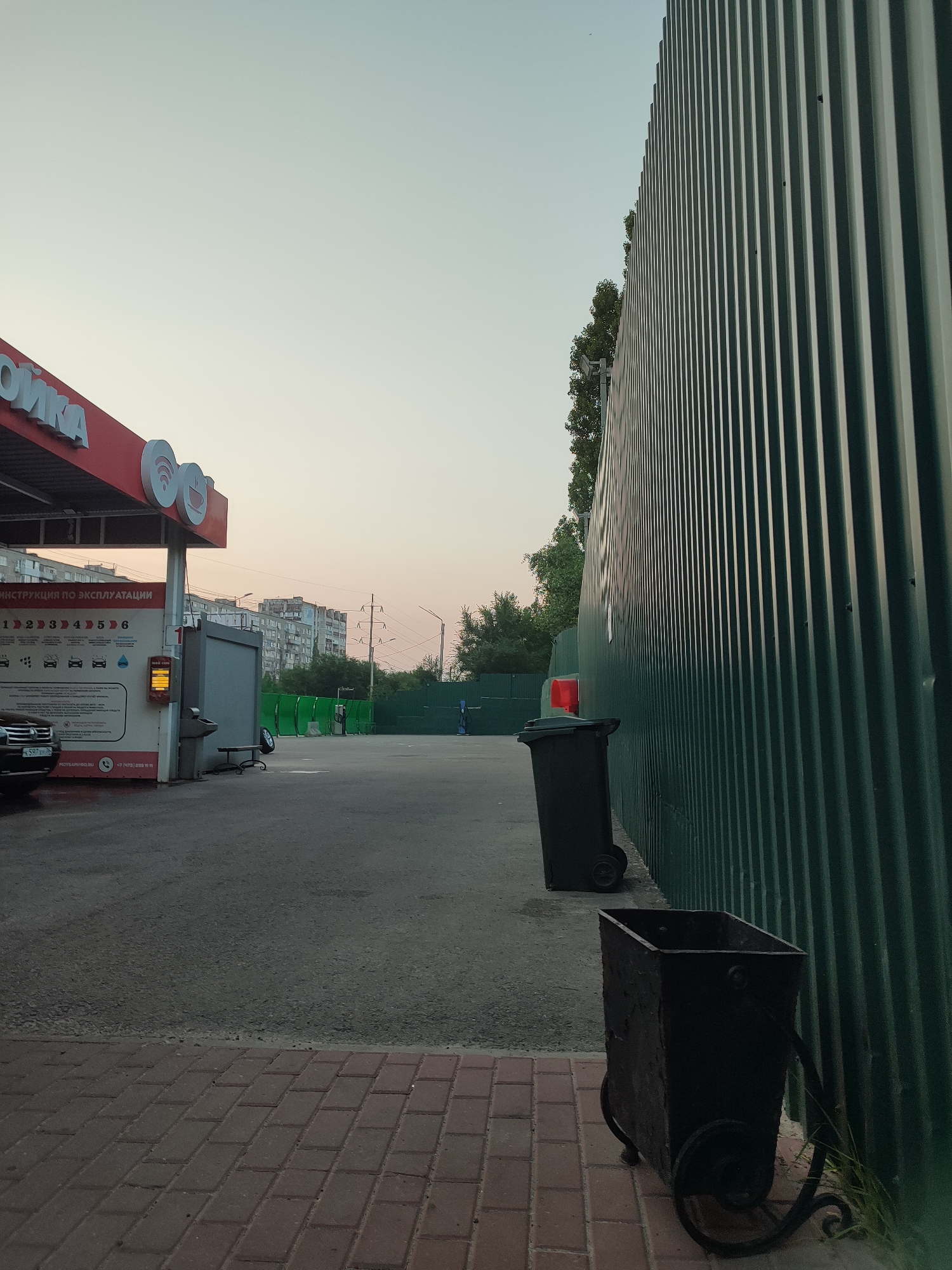 A car wash in Voronezh, Russia, is depicted at dusk, with a clear, pale sky above. A green corrugated metal fence dominates the right side of the frame, casting vertical shadows. In the foreground, a brick walkway leads to a paved area where two dark trash receptacles are situated. To the left, a red structure with white lettering, likely part of the car wash, is visible. This structure has signs with instructions and images of car washing services. Buildings and trees form the background.