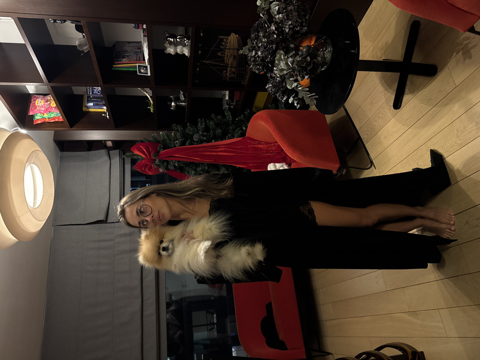 A woman with blonde hair and round glasses poses indoors, holding a fluffy, light brown dog. She is wearing a black outfit. A decorated Christmas tree sits in the background. Behind her is a black bookshelf. A black table with flowers, and red chairs are also visible. The image is rotated, placing the ceiling on the right side. The floor is made of wood.