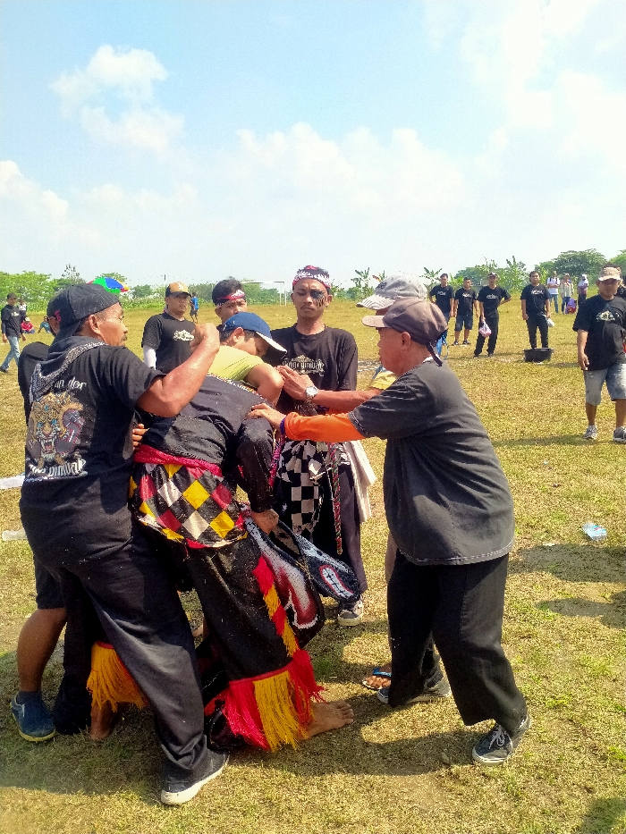 The scene depicts an outdoor gathering on a dry, grassy field in Celep, Indonesia, under a clear blue sky with scattered white clouds. The primary focus is a cluster of approximately six to seven adult males engaged in a dynamic interaction in the foreground.
At the center, one individual is dressed in black clothing, featuring a vibrant red, yellow, and black checkered fabric wrapped around the waist, accented with red and yellow fringed lower leg coverings. This individual is barefoot and appears to be actively moving or being supported. A white-beaded, red-accented object, resembling part of a headdress or an ornamental piece, is visible near their left side.
Surrounding and interacting with this central figure are several men. To the immediate left, a man in a black T-shirt with a graphic design (possibly a tiger) and a black baseball cap firmly grips the central figure's upper body. To the right, another man in a black T-shirt with visible orange long sleeves underneath, and a grey baseball cap, holds the central figure's right arm/shoulder area with both hands, seemingly providing support or guidance. Behind the central figure, a man in a black T-shirt and a blue baseball cap leans into the group, extending an arm. Further back within this central group, a man wearing a black T-shirt, a red and white patterned headband, and dark facial markings observes the interaction, with a white checkered cloth draped over his shoulder. Another man in a black T-shirt and light grey baseball cap stands slightly to the right, also observing.
In the mid-ground and background, numerous other adult males, predominantly in black T-shirts and dark pants or shorts, are dispersed across the field, some observing and others standing casually. A black bucket is visible on the grass in the mid-ground, and a white goalpost frame is discernible in the distant background. A faint white line is visible on the grass in the foreground. The scene is brightly illuminated by direct sunlight, casting distinct shadows.