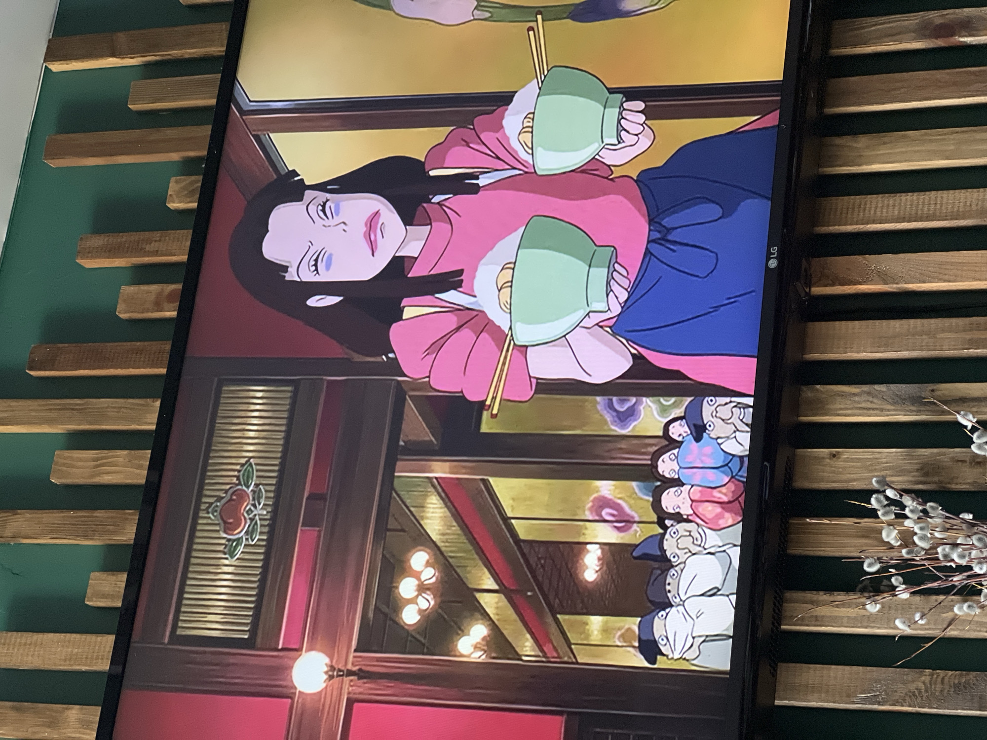 The image shows a television screen displaying a scene from the animated film "Spirited Away" by Studio Ghibli. The character Yubaba is visible, holding two bowls of food with chopsticks. In the background, other characters are present. The television is positioned against a wall with a decorative wooden slat design, and to the right of the screen is a dried flower arrangement. The image doesn't provide any information about the city of Kazan, Russia.