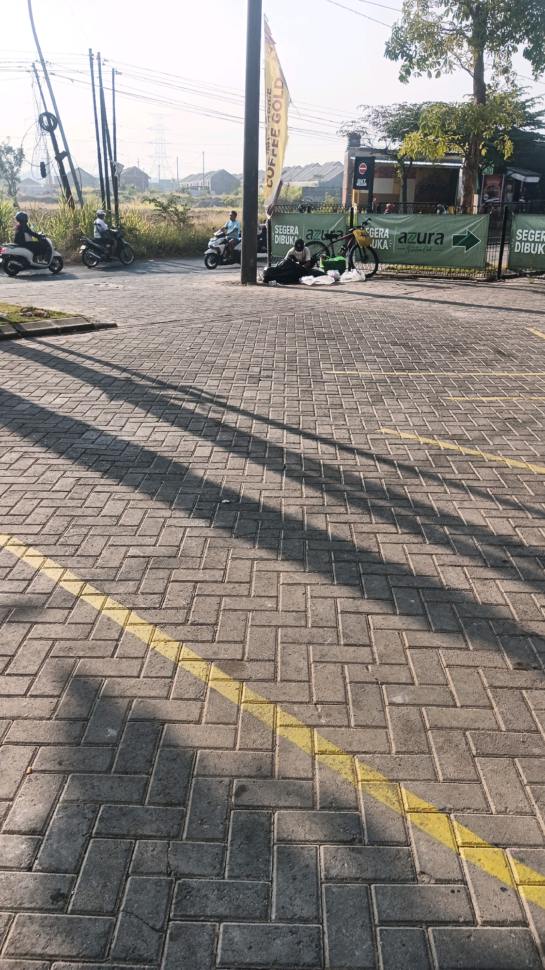 The image depicts a paved area with a herringbone brick pattern, marked by yellow lines. In the background, a street scene unfolds with several motorbikes on a road. Three individuals are on motorbikes, and another is seated on the ground next to a bicycle, seemingly taking a break. A large green banner with the word "azura" and the text "NUTRITION CLUB" is visible. A yellow flag with "COFFEE GOLD" printed on it hangs from a pole. The environment suggests an outdoor setting, possibly near a commercial establishment or rest stop. The lighting indicates it is either early morning or late afternoon, with long shadows cast across the pavement. The location is identified as Mojosarirejo, Indonesia.