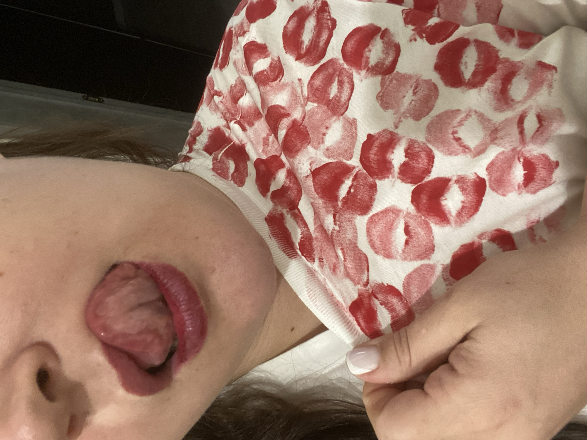 A low-angle shot depicts a person lying down, their head tilted back. The subject has brown hair and fair skin. The person's mouth is open, and their tongue is protruding. They are wearing lipstick. The person is wearing a white shirt with red lipstick print. Their hand is visible, with a fingernail showing a french manicure. A dark object in the background is out of focus, potentially a piece of furniture. No details indicate the location is Sochi, Russia.