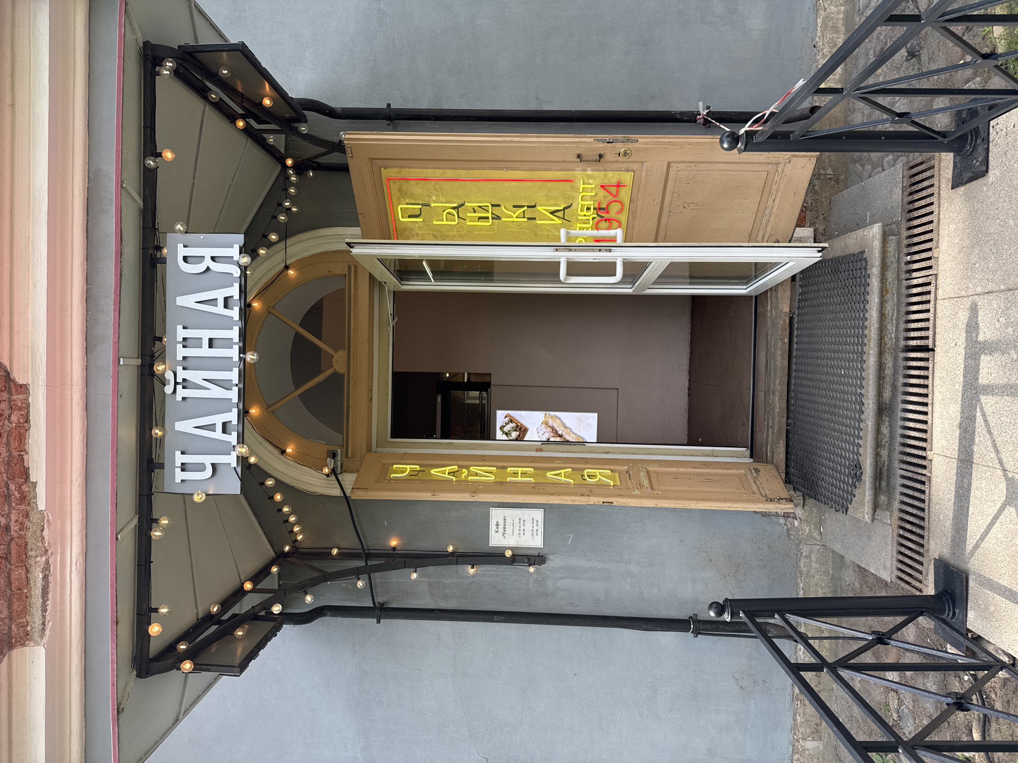 The image captures the street-level entrance of a cafe named "Чайная" (Chaynaya, meaning "Tea Room" or "Tea House") in Saint Petersburg, Russia, bathed in bright daylight. The focal point is a light-colored wooden double door, with one leaf partially open, revealing a dark interior. Above the open door, a yellow neon sign displaying "ЧАЙНАЯ" is visible, appearing mirrored as it's likely placed on the interior side of the glass or door. A smaller red neon sign above it, also mirrored, reads "РЕЦЕПТ" (Retsept, meaning "Recipe") and "054".
To the left of the entrance, a vertical grey sign with prominent white letters again spells out "ЧАЙНАЯ". This sign is positioned beneath a dark metal awning adorned with a string of numerous small, glowing light bulbs, suggesting a warm and inviting atmosphere. The awning extends outwards from the building's facade.
On the grey stucco wall to the right of the door, a small white informational sign states "Кафе «Чайная»" (Cafe "Chaynaya"). Below this, it lists a date range "01.30 по 30.04" (January 30 to April 30), followed by operating hours: "10:00-19:00" and "10:00-20:00", likely indicating different daily or seasonal schedules.
A black metal railing extends from the right side of the entrance along the pavement. A dark, textured doormat is visible on the step just inside the doorway, and a metal drainage grate runs parallel to the building along the sidewalk. The architecture on the far left shows a section of exposed red brickwork and a decorative pink-striped column, characteristic of older urban buildings. No people are visible, and the scene is static, depicting the open, welcoming entrance of the cafe.
