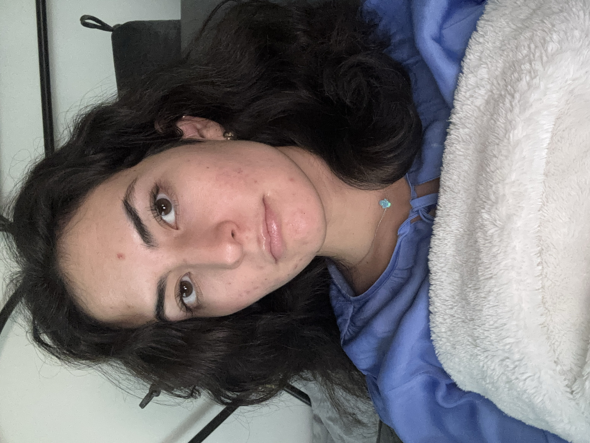 A young woman with dark, wavy hair is lying down, partially covered by a light blue garment and a white, fluffy blanket. She has light skin, brown eyes, and slightly parted lips. A silver necklace with a light blue pendant is visible on her neck. Her right ear is visible with a gold earring. The background includes a black object, possibly a headrest, and a light-colored wall. There is no information available in the image to confirm the location in the city of Puebla, Mexico.