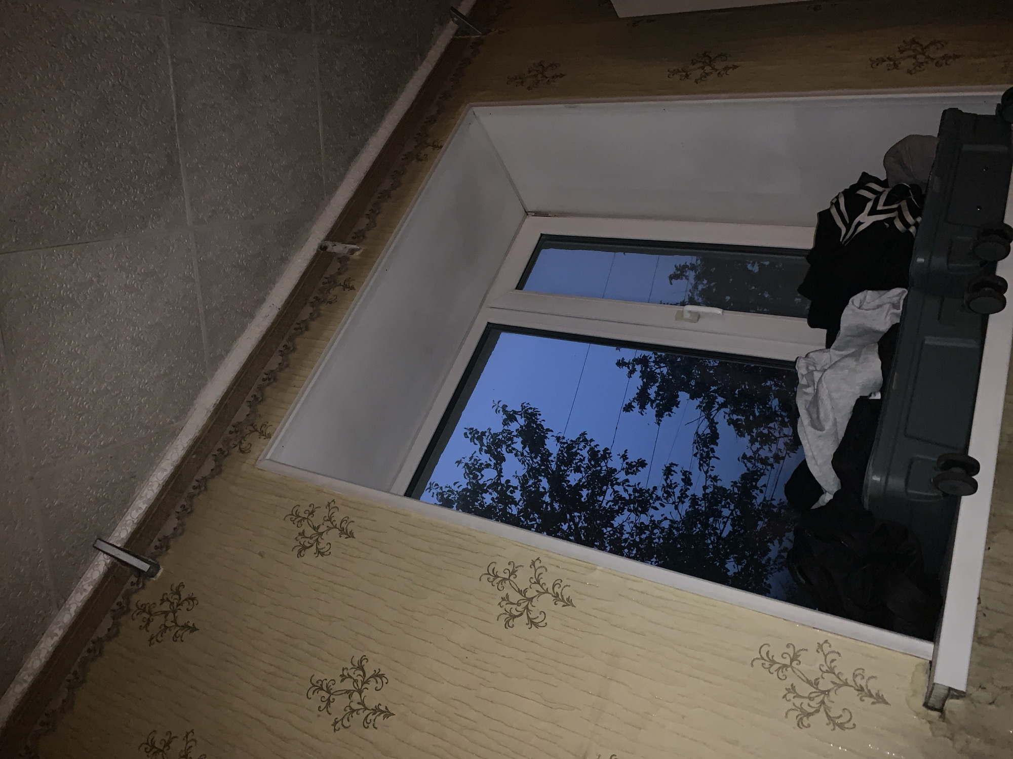 The image shows an indoor scene with a window overlooking trees and the sky at dusk. On the windowsill, a dark green suitcase with wheels is piled with various clothing items, including black, white, and striped garments. The walls are covered in a yellow wallpaper with a repeating ornate pattern. The ceiling appears to be textured and is partially obscured by a white trim. There are no people visible in the image. The overall impression is one of a slightly disheveled domestic space.