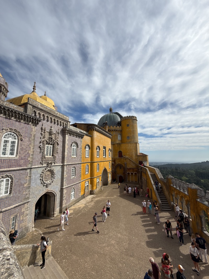 The image captures a scene at the Pena Palace in Sintra, Portugal. The foreground shows a courtyard paved with cobblestones, where a number of tourists are walking and standing. The courtyard is surrounded by architectural structures of the palace. On the left is a building with blue and gray tiles and ornate carvings. Adjacent to it is a yellow building with white-framed windows. A staircase leading to an upper level is visible along the right edge of the courtyard, with people ascending and descending. In the background, beyond the palace walls, lies a view of a forested landscape under a cloudy sky.