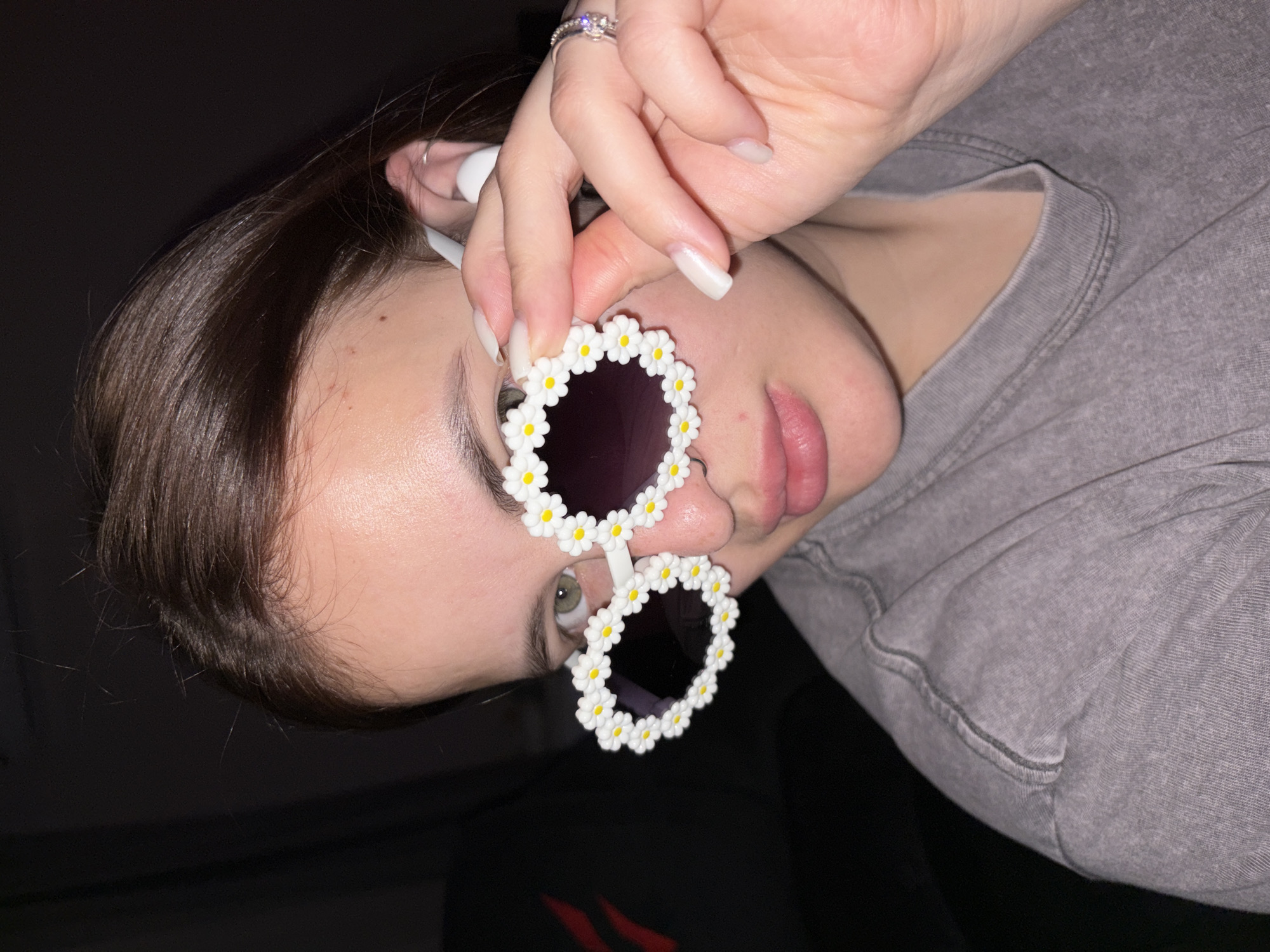 A close-up, vertically oriented portrait shows a young individual, rotated approximately 90 degrees clockwise, primarily focusing on their upper body and face. The individual has medium-dark brown hair and fair skin, and is wearing a light grey crew-neck t-shirt. Their right hand, with neatly manicured, long nails painted a pale neutral color, holds a distinctive pair of white, daisy-shaped sunglasses in front of their face. Each circular lens of the sunglasses is framed by numerous small white petals with yellow centers.
A silver-toned ring with a clear stone is visible on the ring finger of the right hand. Through the bottom lens of the sunglasses, the individual's light-colored eye (appearing green or hazel) is visible, looking directly towards the viewer. A white earbud is seen inserted into their right ear. The lips are slightly parted, showing a natural pink color. The background is mostly dark, with a shadow creating a stark contrast, and the textured grey fabric of the t-shirt fills a significant portion of the right side of the frame. The overall mood conveyed is casual and playful. There is no visible text in the image.