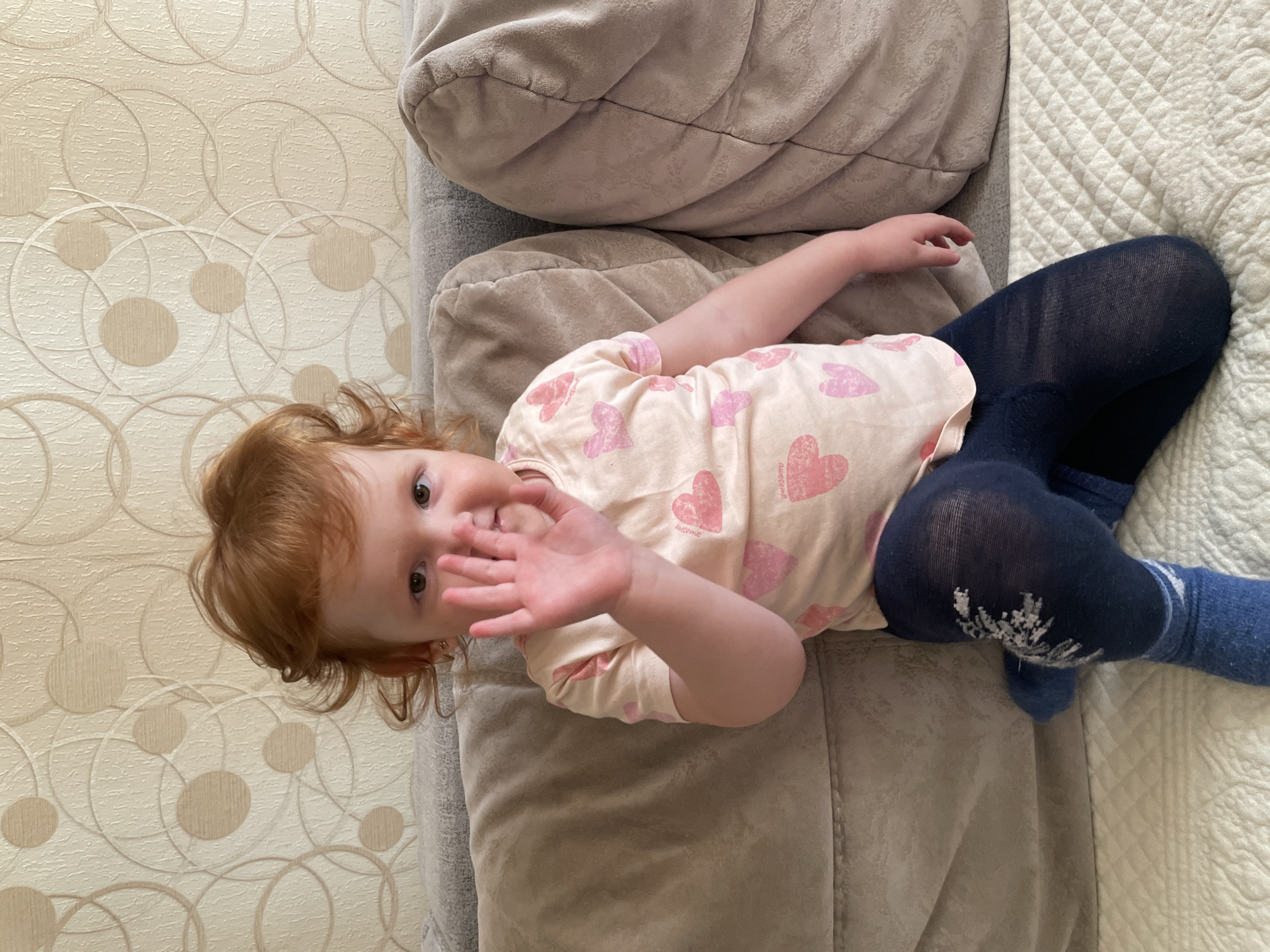 A young child with red hair is lying on a grey upholstered sofa with decorative pillows. She is wearing a light pink long-sleeved shirt with pink hearts on it and dark blue leggings with a design on one leg. The child is waving with her right hand and looking at the camera. The sofa is placed against a wall covered with wallpaper featuring a geometric pattern of circles. A white quilted fabric is also partially visible on the sofa. The location is specified as Кипчак, Russia, but there are no specific elements in the photo confirming the specific location in the city.