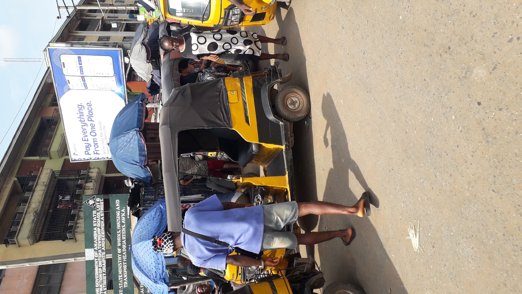 The image depicts a busy street scene in Onitsha, Nigeria, with a strong emphasis on transportation and commercial activity. The focal point is a yellow and black auto-rickshaw, commonly known as a "Keke Napep," parked on a paved road. The auto-rickshaw has a black canopy and black seats, and a "DELUXE" label is visible on its side.
Several people are present. A man wearing a blue shirt, denim shorts, and flip-flops stands near the auto-rickshaw, facing away from the camera. He has a black bag slung over his shoulder. A woman in a black and white polka dot dress and sandals walks towards the right side of the frame. Another person in a striped shirt and red shorts is visible inside the auto-rickshaw. Two blue umbrellas are set up near the auto-rickshaw, likely for shade.
In the background, buildings with multiple stories and balconies are visible. A prominent billboard displaying an advertisement for "Palipay" with the slogan "Pay Everything. From One Place" is mounted on one of the buildings. Signs with text, including one that reads "THE GOVERNMENT OF ANAMBRA STATE" and another with "TRANSPORT HEADQUARTERS, AWKA," are also present, providing contextual clues about the location within Onitsha. The ground is a mixture of asphalt and concrete, with shadows cast by the vehicles and people, indicating a bright, sunny day. Scattered debris, including what appears to be dried plant material, is on the ground. The scene captures the vibrant, everyday life of a Nigerian city.