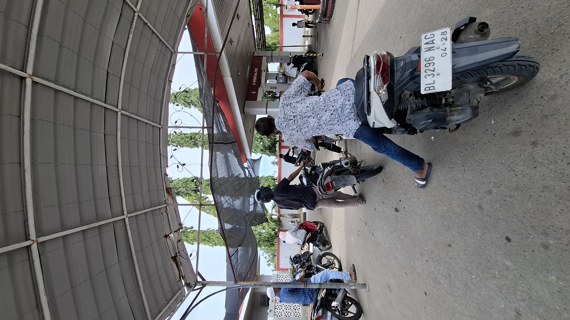 The image depicts a daytime scene at a Pertamina gas station, identified by the "PERTAMINA" signage. The location is specified as Keudebayu, Indonesia, based on context from the user's prompt. Several people are present, most of them on or next to motorcycles, seemingly waiting in line to purchase fuel. One person is wearing a patterned shirt and jeans, another a black t-shirt and pants, and a third a blue checkered shirt and light blue pants. A woman in a striped shirt and headscarf is also visible on a scooter. The gas station has a covered area with a corrugated metal roof supported by metal beams. Behind the covered area, a red and white structure houses the fuel pumps, with the number "4" and "PERTALITE" displayed. Tall, thin trees with dense foliage are visible in the background, along with a pale blue sky. The ground is asphalt. One motorcycle in the foreground has a license plate that reads "BL 3296 NAG 04.28". The overall impression is of a routine activity at a local gas station.