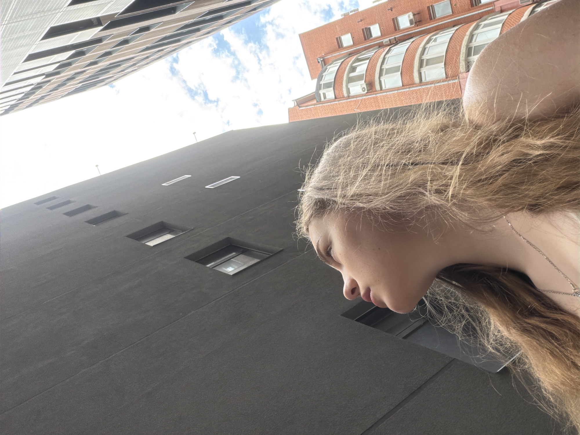 The image is an upward-angle shot of a young woman in Dnipro, Ukraine. She is looking upwards, with her profile visible against the sky and buildings. The woman has long, wavy blonde hair. She is wearing a silver necklace. In the background, there are modern and older brick buildings. The sky is partly cloudy. The perspective suggests the photo was taken in a narrow street or between buildings.