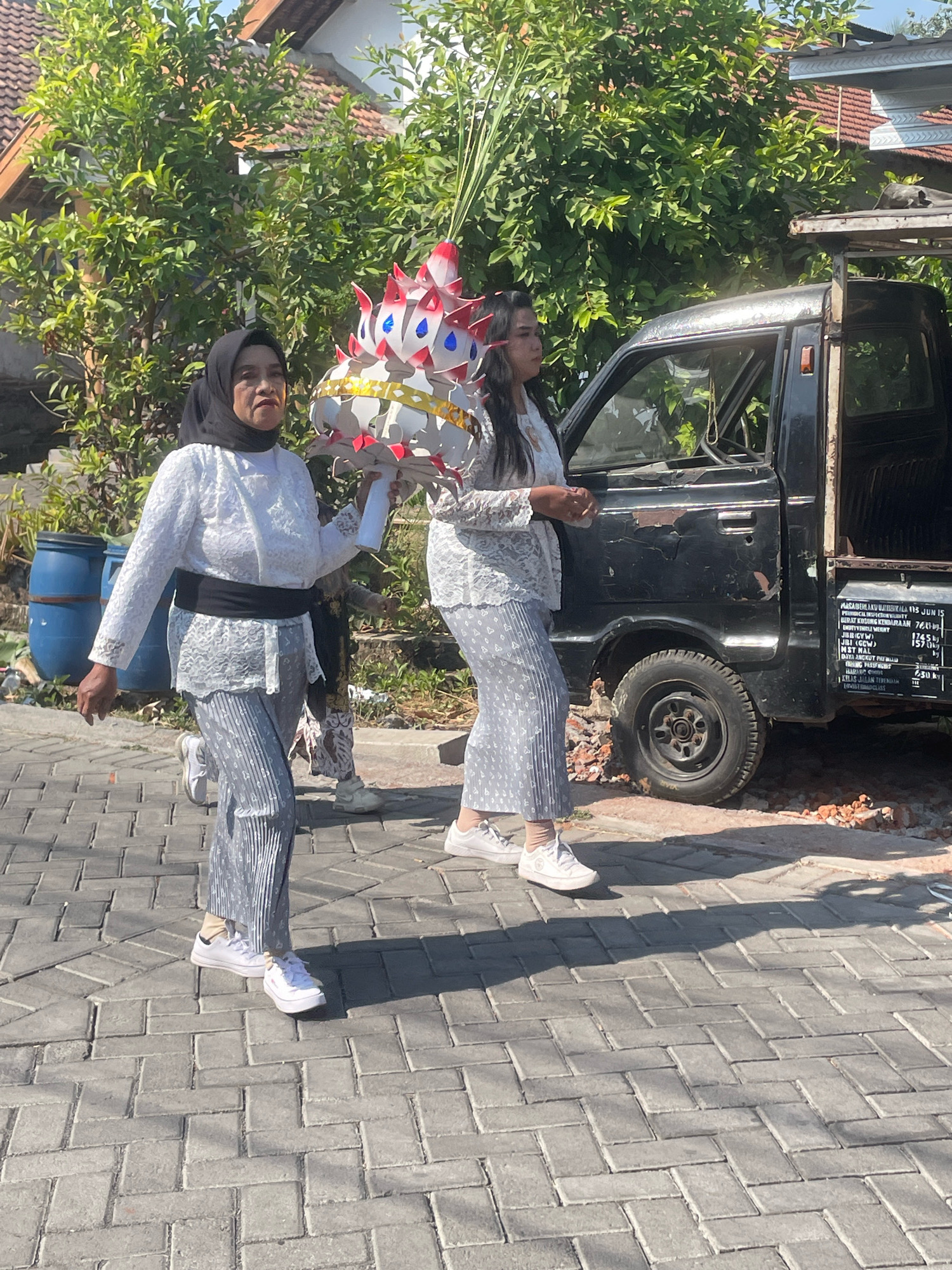 In Kebon Waris, Indonesia, two women in traditional attire walk on a paved street. The woman on the left wears a white lace top, a black sash, and patterned pants, holding a festive, conical ornament. The woman to her right wears a similar white lace top and patterned pants. A black pickup truck is parked next to them. In the background, a house with a tiled roof and lush green trees are visible. The scene is illuminated by bright sunlight, casting shadows on the ground.