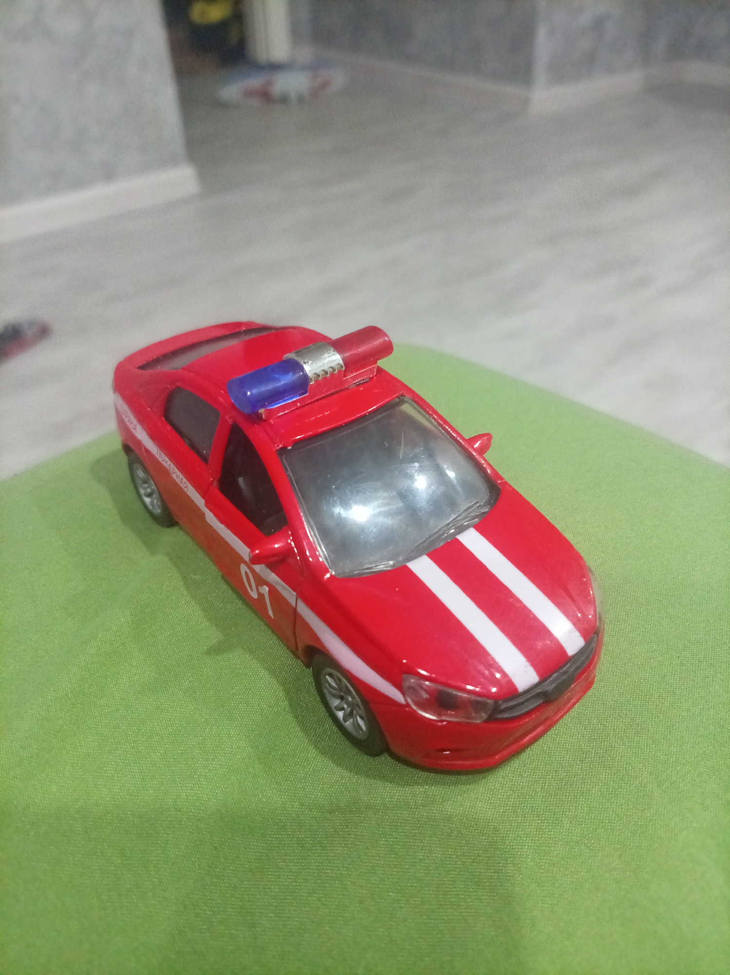 A close-up, slightly elevated shot shows a red toy emergency vehicle with white racing stripes on its hood. A blue and red siren light bar is mounted on the roof. The toy car has "01" in white numerals on its side. The vehicle rests on a textured green surface, possibly fabric. In the background, a blurry grey wall and floor suggest an indoor setting. The provided hints show details of the car and the immediate background. The location is in the city of Oktyabrsk, Russia.