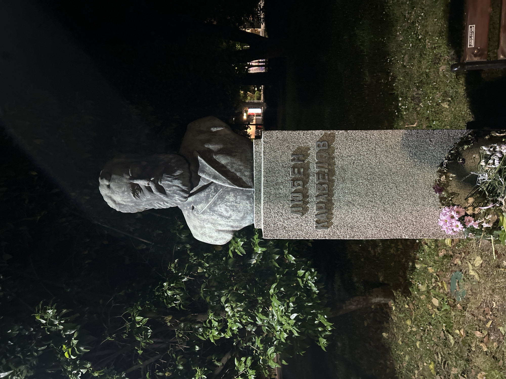 A sideways nighttime shot depicts a monument featuring a gray stone bust on the left and a rectangular stone plaque on the right. The plaque bears the Cyrillic inscription "ИВАН ВАЗОВ" in vertical, blocky letters. Fresh flowers are arranged at the base of the plaque. The statue and plaque appear to be located in a garden or park setting, with foliage visible around the monument. In the blurred background, faint lighting suggests the presence of buildings and possibly street lamps, indicating the monument is in an urban environment, likely Burgas, Bulgaria.