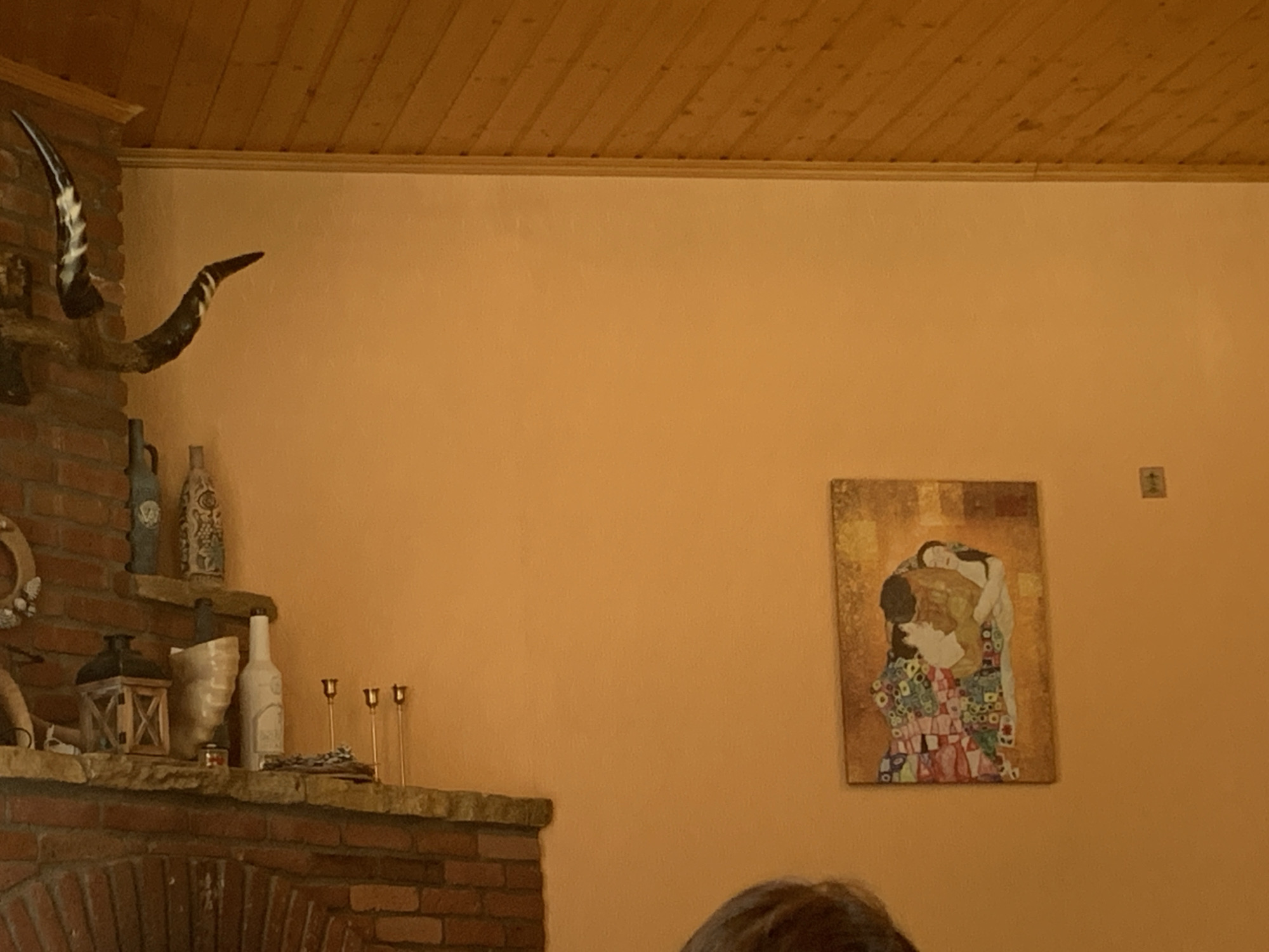 The image shows an indoor scene with an orange wall and a wooden ceiling. On the left side of the wall, a large horn is mounted. Below the horn, a brick fireplace with a stone mantelpiece is visible. The mantelpiece holds several decorative items, including a lantern, vases, and bottles. To the right of the fireplace, a painting is hanging on the wall. The painting depicts two figures embracing, rendered in a vibrant, mosaic-like style. A small, dark square object is also visible on the wall to the right of the painting. The bottom of the image shows the top of a person's head, suggesting they are seated or standing in front of the scene. The overall ambiance is warm and rustic.