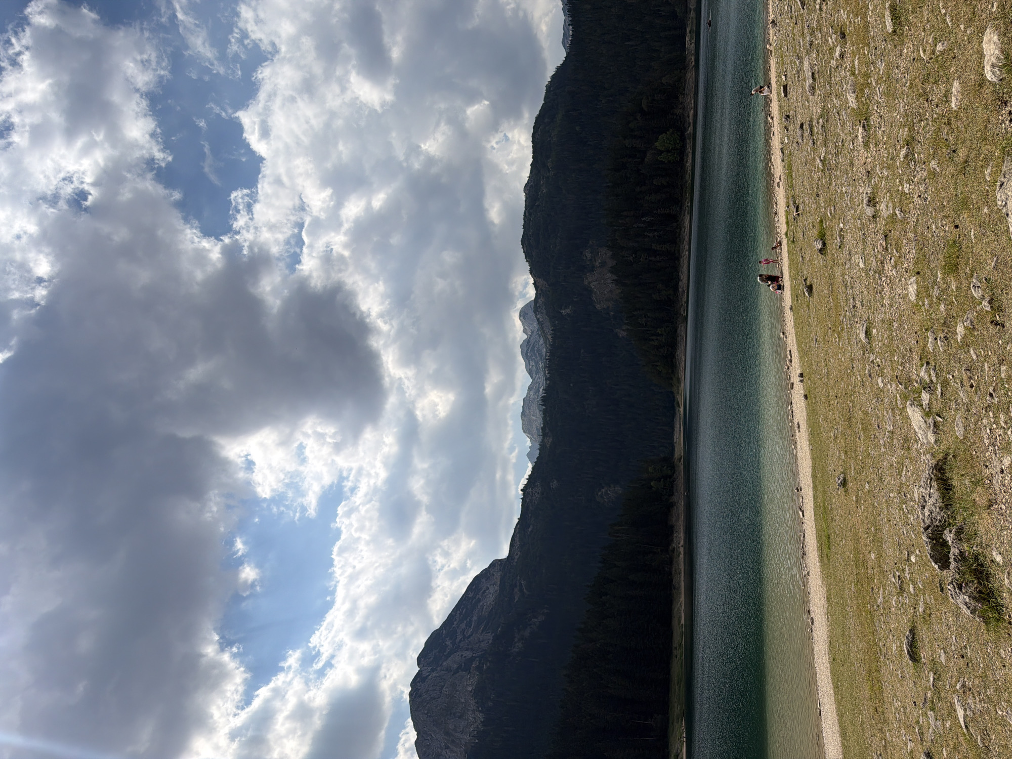 The image shows a serene lake scene with a backdrop of mountains and a cloudy sky. In the foreground, a rocky, grassy shore slopes gently towards the water. The lake itself is a deep, rich teal color, with lighter, sandy shallows near the shore. A dense forest of dark green pine trees covers the lower slopes of the mountains, which rise up dramatically behind the trees. The peaks of the mountains are rugged and rocky, with some areas appearing snow-dusted. The sky is a mix of dramatic, dark grey clouds and patches of bright blue sky, with sunlight breaking through in places, casting dramatic shadows. On the shore of the lake, a few individuals are visible. On the left side of the frame, a small group of people are gathered near the water's edge. Further to the right, a solitary figure is seated on the shore. The scene is peaceful and natural, suggesting a location in a mountainous, natural park area. This scene is reminiscent of the natural beauty found in the vicinity of Pitomine, Montenegro, known for its stunning mountain lakes and forests.