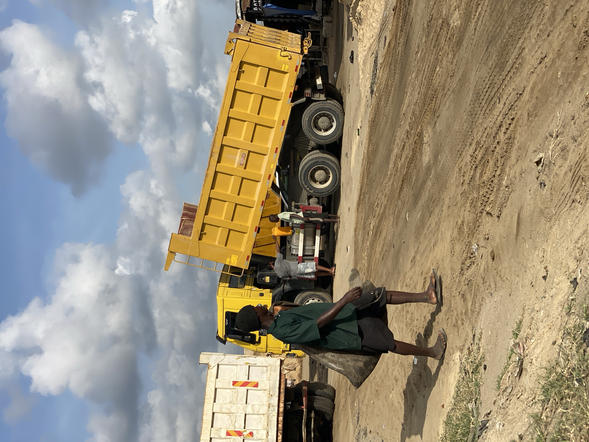 The image shows a man walking on a dirt road in Ado, Nigeria, carrying a bag on his shoulder and holding what appears to be a hat. He is wearing a green shirt, black shorts, and sandals. Behind him are two yellow dump trucks, one with its bed raised. Two other men are visible near the raised truck. The road is dirt with tire tracks, and the background shows a partly cloudy sky and some vegetation.