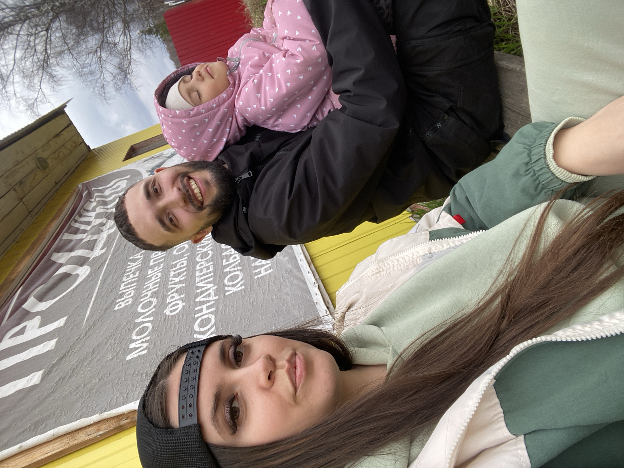 The image captures a selfie of a young woman with long brown hair and a black baseball cap. Behind her, a man with a beard smiles, holding a sleeping child wearing a pink jacket with white hearts. They are outdoors, with a yellow building and a sign in Russian visible in the background. The sign is advertising various food products including baked goods, dairy products, fruits, sweets, and sausage. The location is the city of Zhigalki, Russia.