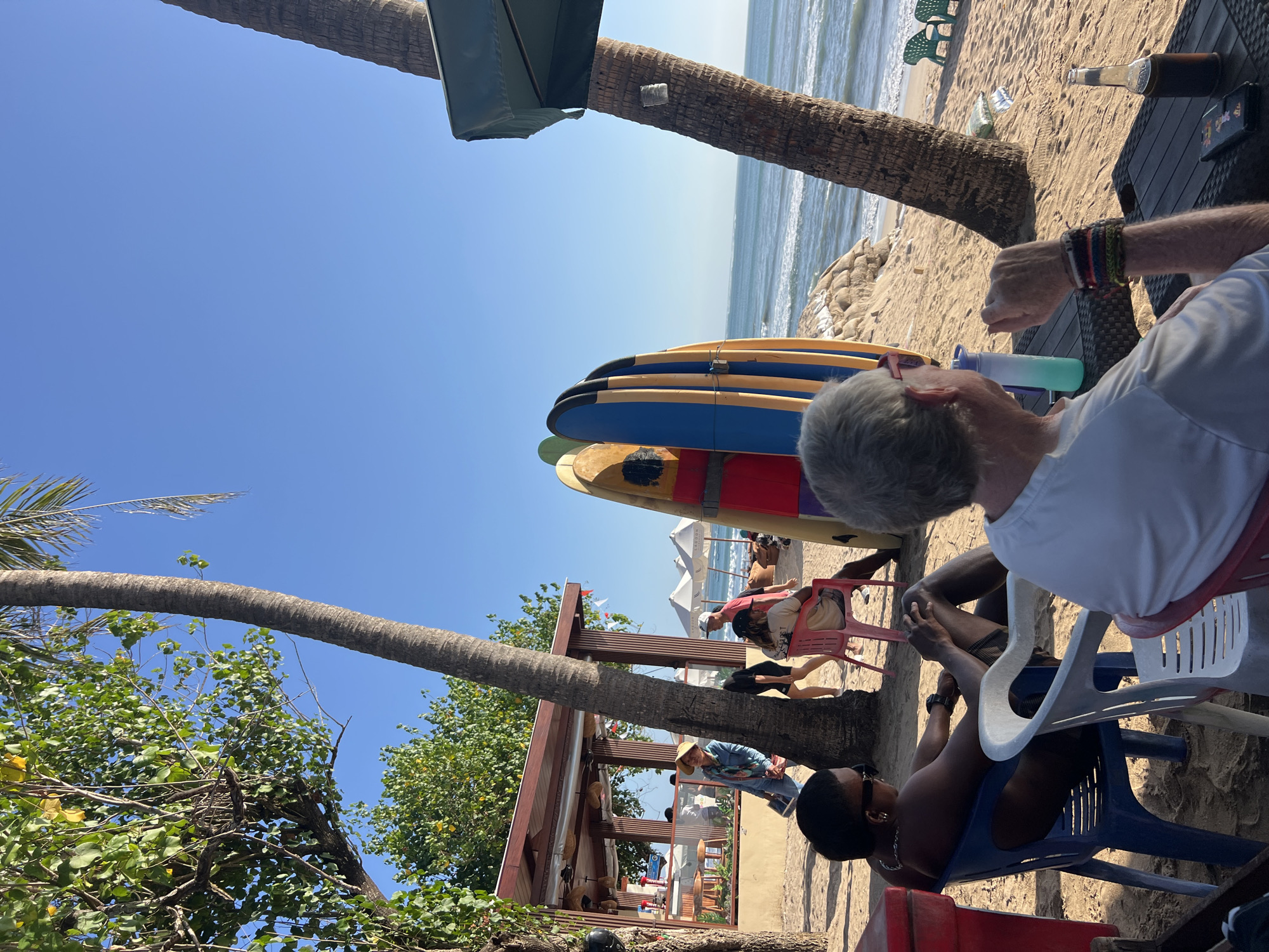 A group of people are gathered on a sandy beach in Kuta, Indonesia, near a beachfront establishment. In the foreground, two individuals are seated in chairs facing away from the camera. To the left, a person wearing sunglasses and a chain is seated in a blue plastic chair. To the right, another person, partially visible, sits in a white chair. A table between them has a bottle and a phone on it.
In the mid-ground, several surfboards are propped up vertically. Behind the surfboards, a person in a red top and white hat is walking, and another person is seated in a pink chair. Further back, a covered structure with tables and chairs suggests a seating area for the establishment. People are seen walking and interacting in this area.
The background features the ocean with gentle waves and a clear blue sky. Palm trees are visible throughout the scene, some with leaves and others more bare. Beach umbrellas are also scattered on the sand. The overall atmosphere appears relaxed and casual, typical of a beachside location.