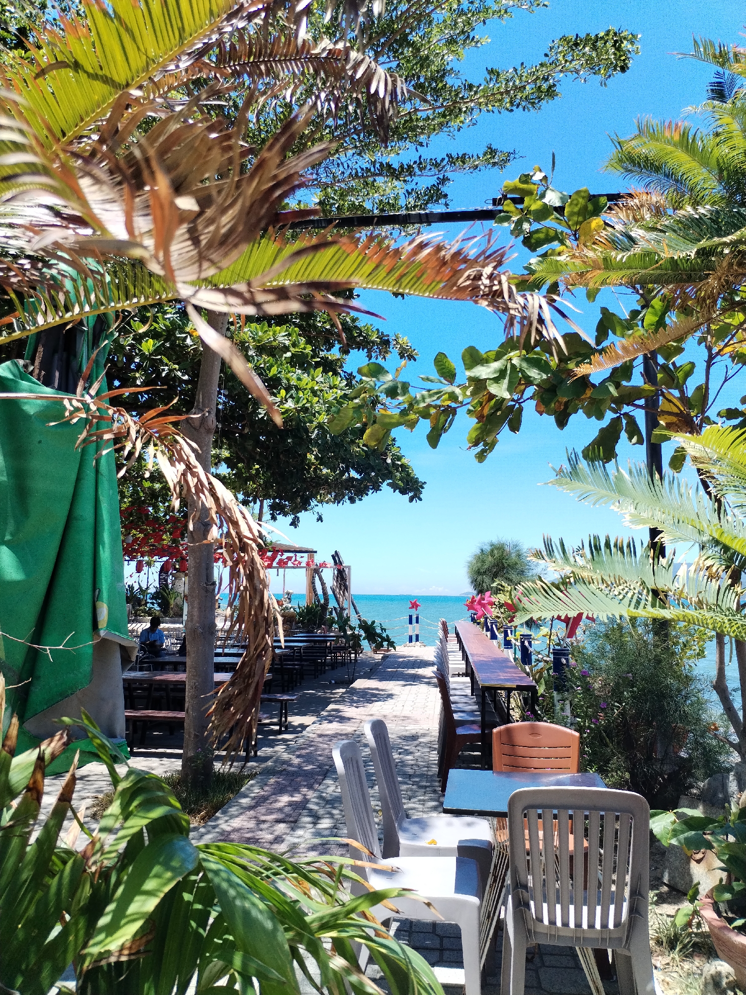 This image shows a perspective of a restaurant or cafe extending towards the coast of Nha Trang Ward, Vietnam. The foreground contains an outdoor dining area with plastic chairs and tables. Foliage, including palm trees and other greenery, frames the view. In the distance, a boardwalk extends to the water. Patrons are seated at the tables. The sky is bright blue.