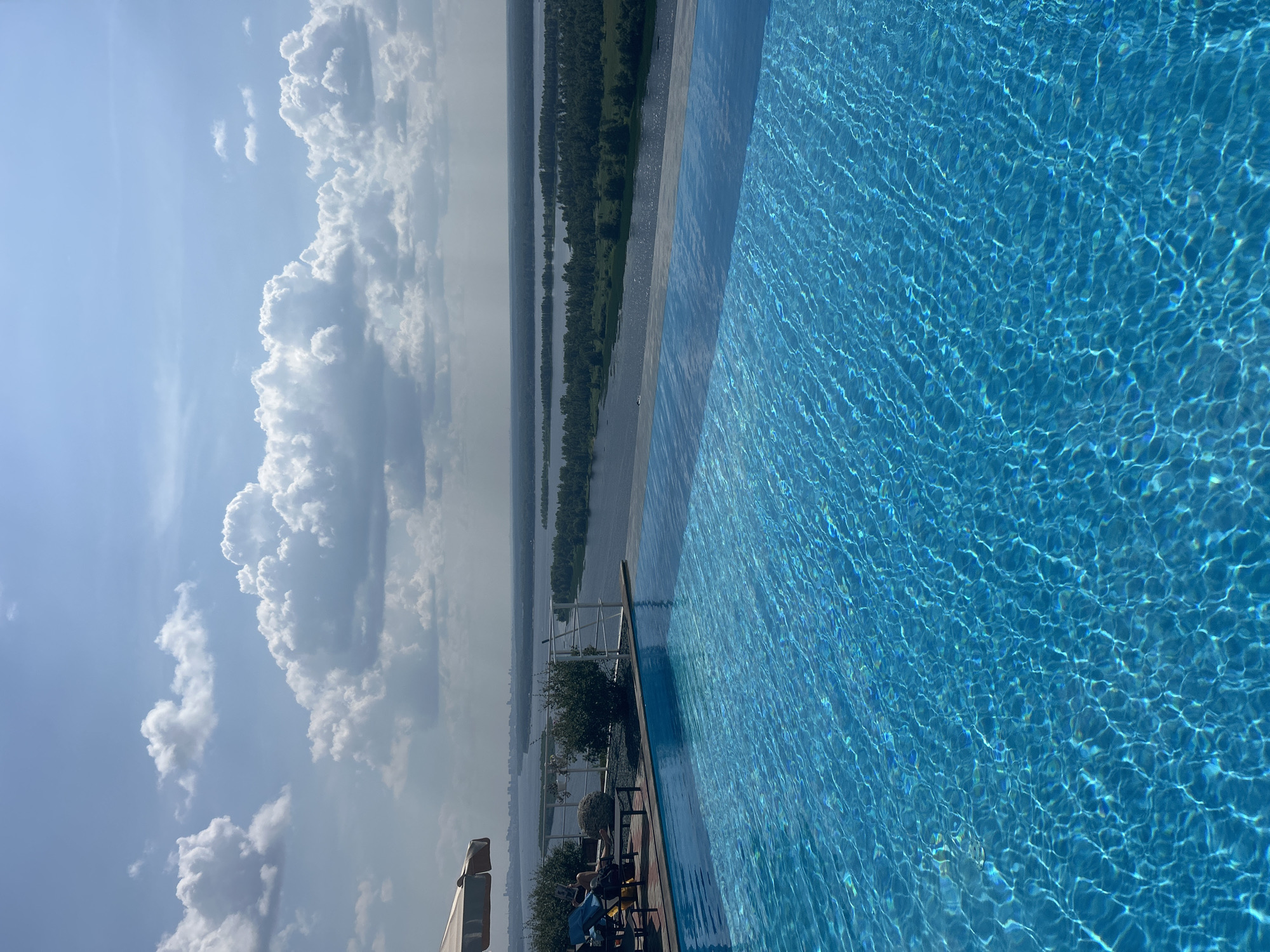 An infinity pool is filled with bright blue water that reflects the sunny sky. The pool is on a terrace that overlooks a wide river and a tree-lined bank. In the distance, a city skyline is visible. A white railing separates the pool area from the view. There are lounge chairs, a table, and an umbrella on the terrace. Two people are seated in lounge chairs. The sky is filled with fluffy white clouds. The location is the city of Novokuybyshevsk, Russia.