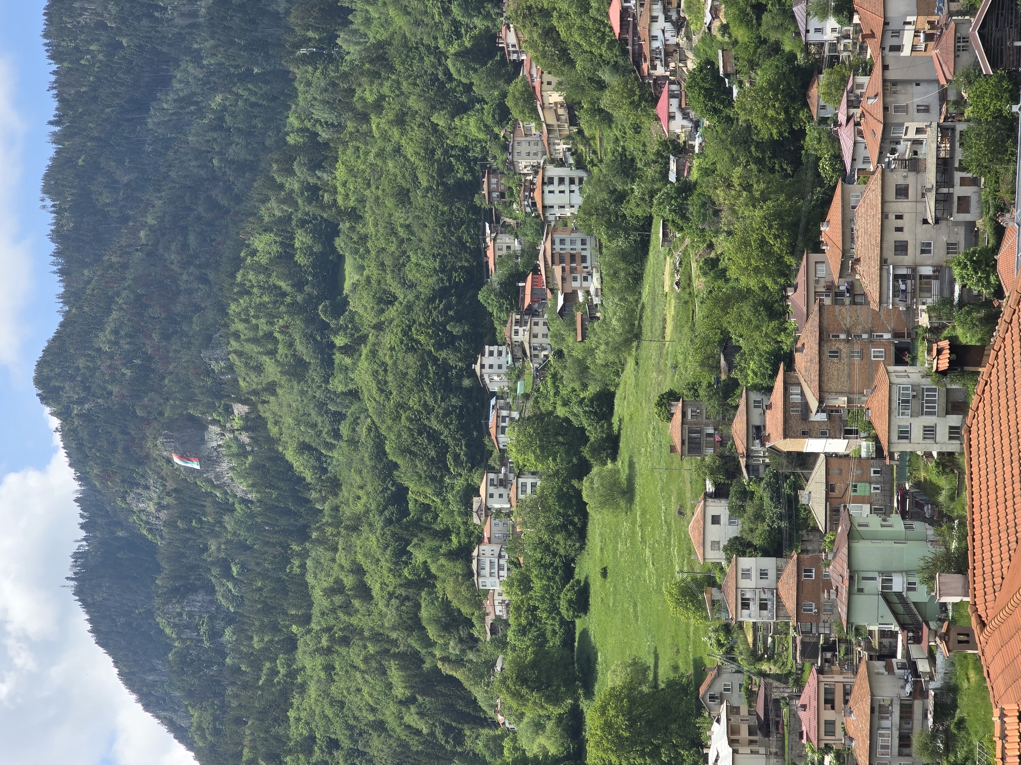The image presents a panoramic view of a hillside town nestled against a densely forested mountain, under a bright blue sky with scattered white clouds. The perspective appears to be from a higher vantage point, looking down on the settlement.
The left side of the frame is dominated by a majestic, tree-covered mountain. Its slopes are blanketed in a thick, dark green forest. High up on a rocky section of this mountain, a prominent flag is visible, displaying horizontal stripes of white, green, and red.
The right side of the image features a residential area. Numerous houses, characterized by their reddish or orange-tiled roofs, are built across the verdant slopes. The buildings vary in size and architectural style, often multi-storied, and are interspersed with patches of bright green grass and smaller trees, blending into the natural landscape. The arrangement of the houses suggests they follow the contours of the hill.
The scene depicts a peaceful, sunny daytime setting. No human activity or specific events are visible, contributing to the tranquil atmosphere. The light is clear, casting gentle shadows that define the landscape. The combination of dense forest, hillside architecture, and the distinct flag suggests a location in a mountainous region, possibly in Central or Eastern Europe. There is no visible text other than the colors of the flag.