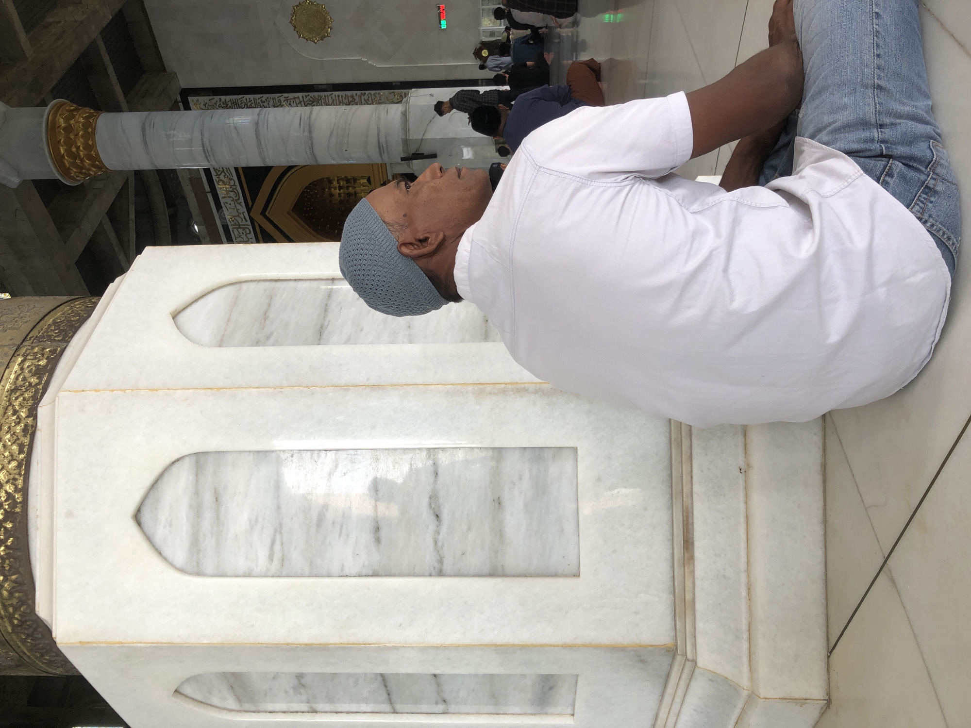 A man wearing a gray skullcap and a white shirt and blue jeans is seated beside a white marble structure inside a building that may be a mosque. Other people are visible further into the building, kneeling on the floor. A pillar with a gold and white base and body is on the left of the man. The location may be Lhokseumawe, Indonesia.