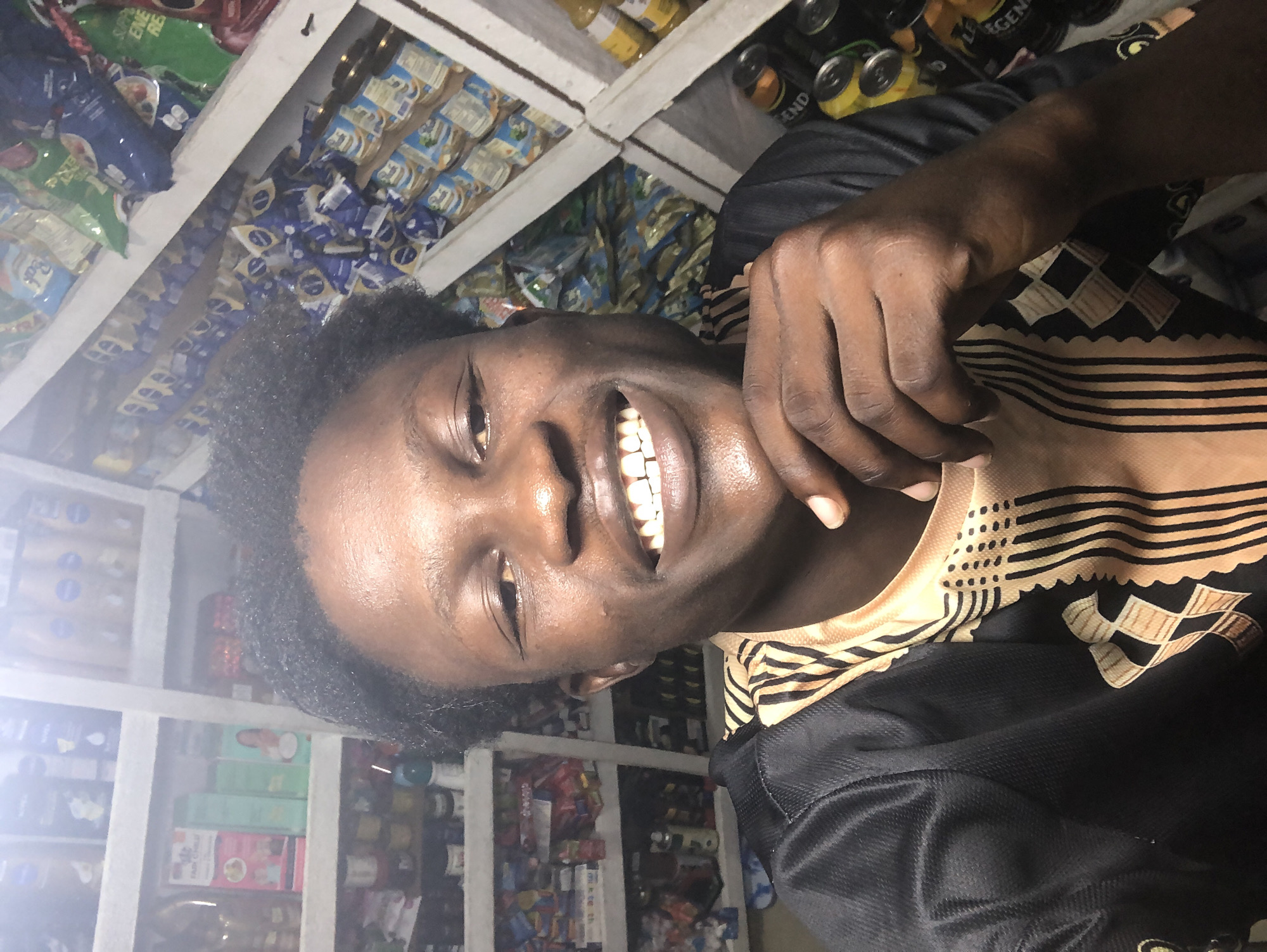 A young Black male is taking a selfie in a shop in Ado, Nigeria. He is wearing a black and gold patterned shirt. The background shows shelves stocked with various products, including food items, toiletries, and drinks. The lighting appears to be artificial, likely from within the shop. The man is smiling widely, with his right hand resting on his chin. The shelves are made of wood and are filled with colorful packaging.