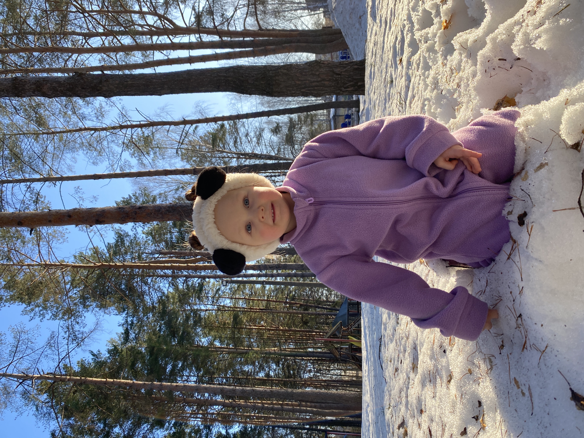 A young child in a purple snowsuit and panda-ear hat is playing in the snow in a wooded area. The child is partially submerged in the snow, with one hand reaching out to touch the snow. Tall pine trees are visible in the background, reaching toward a blue sky. A playground is visible in the background. The location is Chaikovsky, Russia.