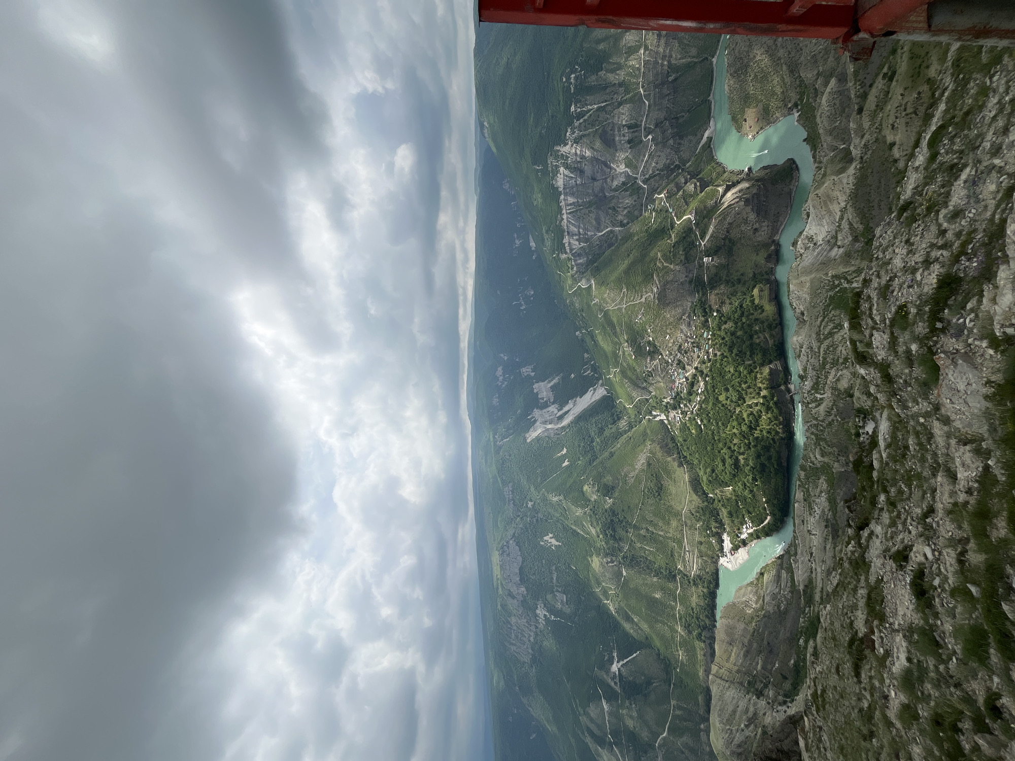 A high-angle, eye-level view captures the Sulak Canyon near the village of Dubki in Russia. A river with a turquoise hue snakes through the canyon floor, flanked by steep, rocky cliffs and verdant vegetation. The sky is predominantly overcast, with varying shades of gray clouds casting a diffuse light over the landscape. A portion of a red structure, possibly part of an aircraft or vantage point, is visible in the upper right corner of the frame.