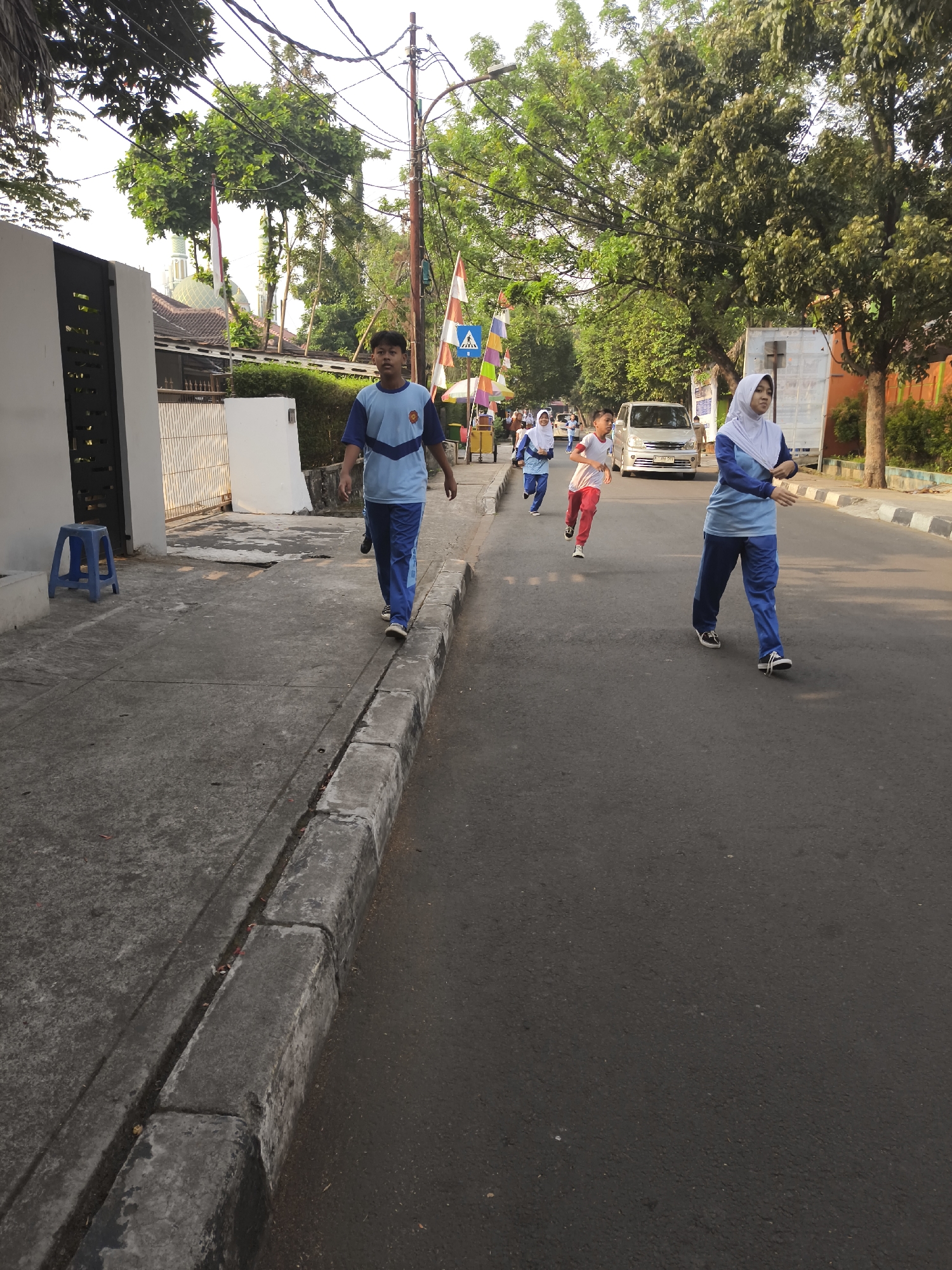In South Jakarta, Indonesia, a group of children in school uniforms are participating in a fun run. The image captures them on a paved road, with a sidewalk on the left. The children are dressed in light blue shirts with darker blue trim and matching blue pants. Some are wearing white hijabs. They are running in the same direction as the camera. In the background, trees line the street, and a silver van is visible. Indonesian flags are displayed on poles along the road, suggesting a national celebration. The foreground shows the gray concrete sidewalk and the asphalt road. A blue plastic stool sits on the sidewalk near a white building with a black gate. The lighting indicates it is daytime, likely morning or late afternoon, with the sun casting shadows.
