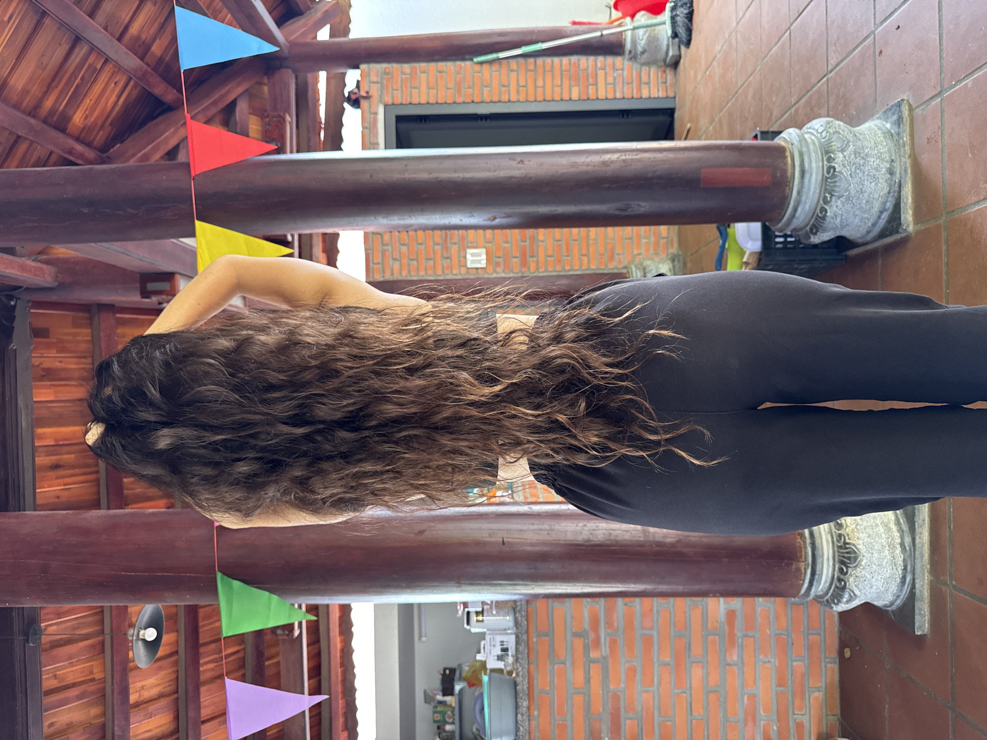 A female subject, viewed from behind, stands centrally in a covered outdoor or semi-open architectural space in Mũi Né, Vietnam. Her long, dark brown, wavy hair extends to her lower back, and her hands are raised, touching the back of her head. She wears a dark top, likely a sports bra, and loose-fitting black pants. The setting features prominent dark brown, cylindrical wooden pillars with ornate grey stone bases, supporting a high wooden roof with exposed rafters and reddish-brown planks. The floor is tiled with reddish-brown square tiles. A string of colorful triangular bunting (purple, green, yellow, red, blue) is strung between the pillars. In the left background, a counter with a grey basin, bottles, and white boxes suggests a utility or kitchen area, backed by a reddish-orange brick wall. In the right background, another reddish-orange brick wall features a dark doorway, and a green-handled broom leans against a pillar, with a red container visible nearby on the ground. A dark metal pendant light fixture with an exposed bulb hangs from the wooden ceiling in the upper left quadrant.