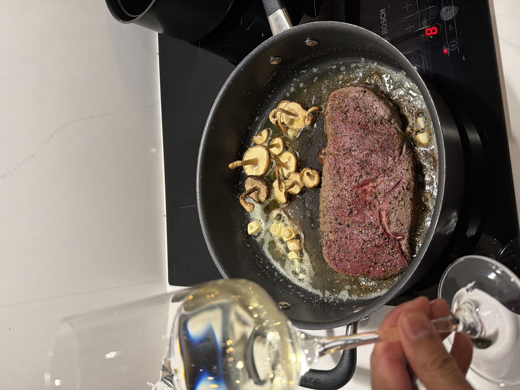 A right hand, holding the clear stem of a wine glass filled with a light, translucent liquid, occupies the lower-left foreground. The glass exhibits multiple bright light reflections and refractions.
The primary focus is a dark grey frying pan positioned centrally on a black Bosch induction hob. Within the pan, a thick, raw-edge steak, visibly seasoned with coarse black pepper and other dark spices, is searing in bubbling, melted fat. The exposed surface of the steak displays a reddish-pink hue, indicative of a medium-rare internal temperature. Several small, light-brown mushrooms with discernible stems, alongside multiple peeled and sliced garlic cloves, are also cooking in the fat surrounding the steak.
The Bosch induction hob features the brand name "BOSCH" in its upper right quadrant and displays the red digital numeral "8," signifying a heat setting. Various touch control symbols are visible on the hob's surface. The background consists of a smooth, light-colored kitchen countertop, possibly white or light grey, extending vertically. A dark cylindrical object, potentially a pot or small appliance, is partially visible on the far right edge of the counter. The environment is a modern kitchen. The scene is located in the city of London, United Kingdom.