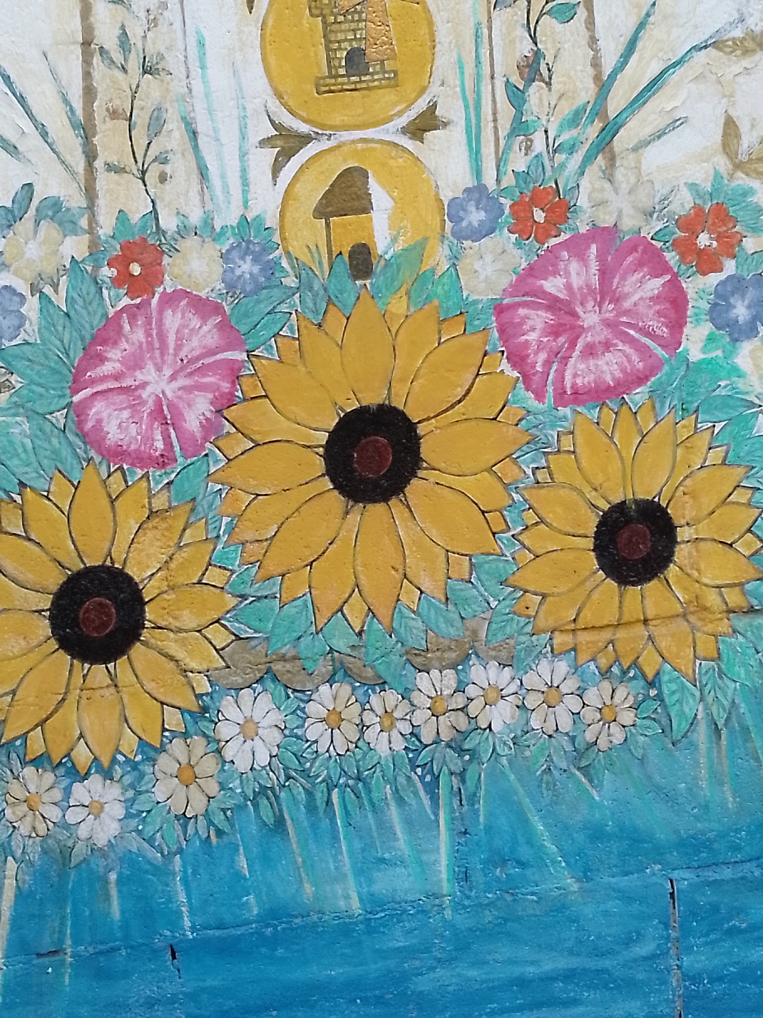 The image shows a section of painted mural in Porlamar, Venezuela. The mural features several stylized sunflowers with yellow petals and dark brown centers, interspersed with pink and blue flowers. A row of small white daisies borders the bottom of the sunflowers. The background includes light teal foliage and blue vertical strokes, possibly representing stems or water. At the top of the mural, two yellow circles each containing a simple depiction of a building. The paint is applied to a textured surface, suggesting a wall or similar structure.