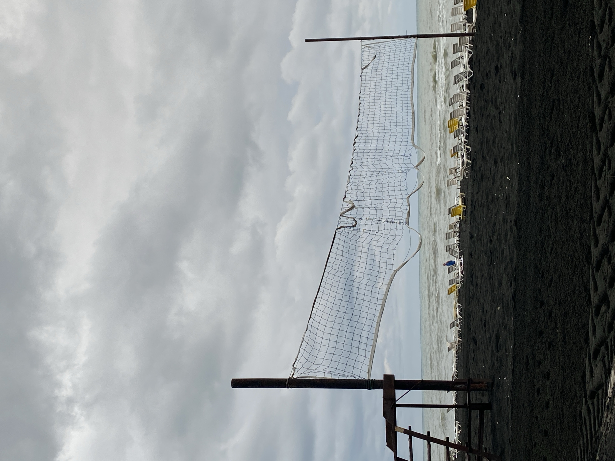 The image depicts a black sand beach in Ureki, Georgia, under a cloudy sky. A volleyball net stretches across the frame, supported by two wooden poles. To the left, a wooden structure with a ladder is attached to one of the poles. In the background, rows of empty lounge chairs are arranged on the sand, facing the wavy sea. A single person in a blue top is visible in the distance, standing near the lounge chairs. The sand shows tire tracks, suggesting vehicle activity. The overall atmosphere is somber due to the overcast weather and the absence of people actively using the beach.
